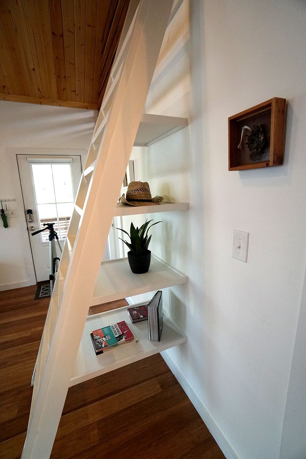 White ladder with shelves against a white wall. Shelves hold plant, hat, books. Wooden floor.