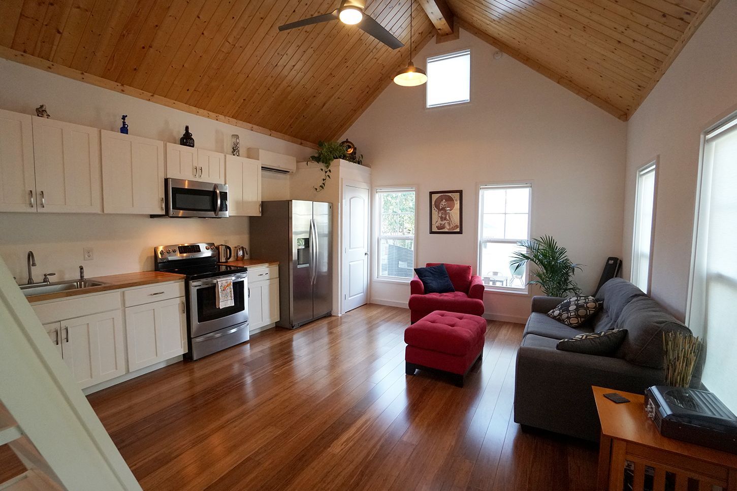 Cozy living space with kitchen area, wood floors, and vaulted wood ceiling; red ottoman, gray couch.