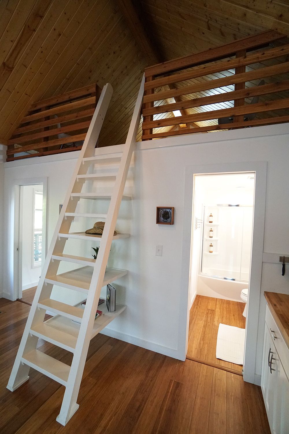 White ladder leads to a loft in a small room with wood floors. A bathroom is visible in the doorway.