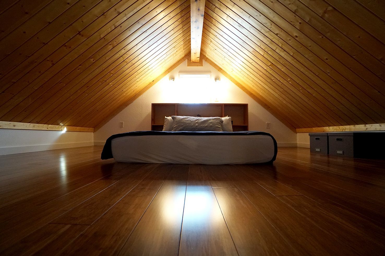 Bedroom in a finished attic with wood floors and vaulted ceiling, bed centered, light coming from window.