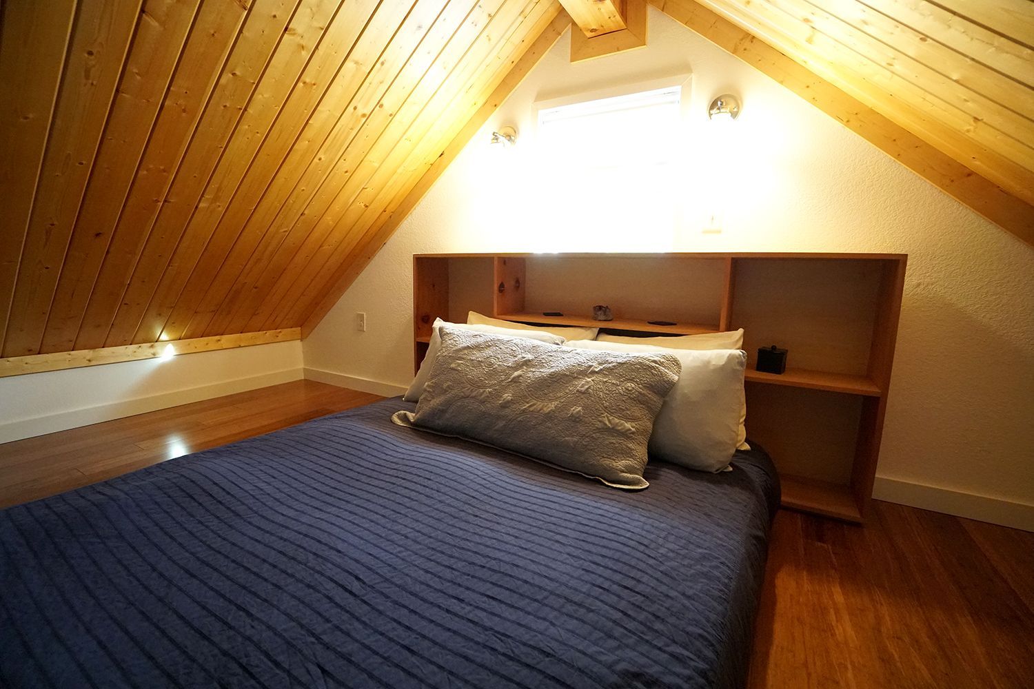 Bedroom with wooden sloped ceiling and floor; bed with blue comforter, headboard with shelves.