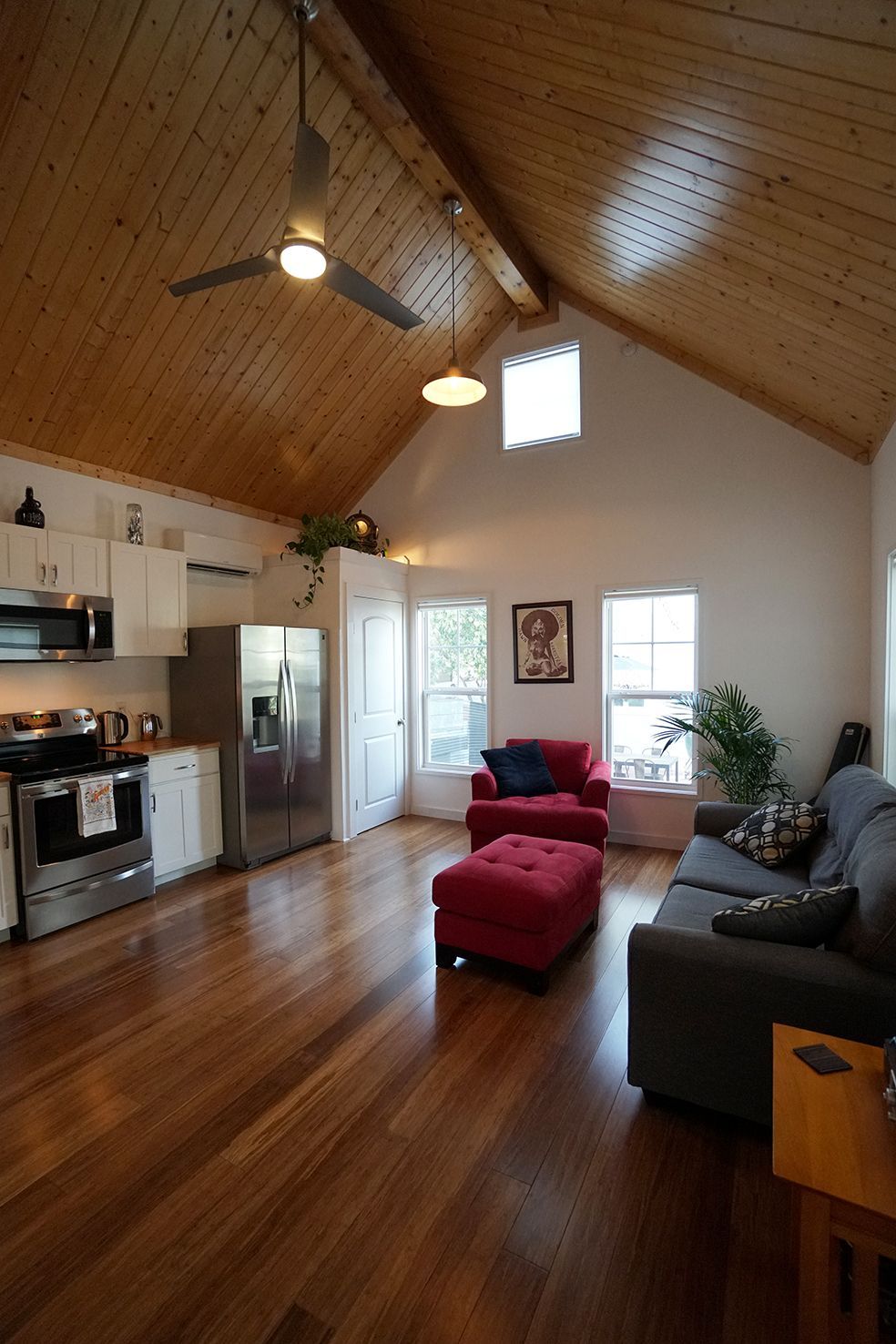 Living room with vaulted wood ceiling, kitchen area, red seating, and wood flooring.