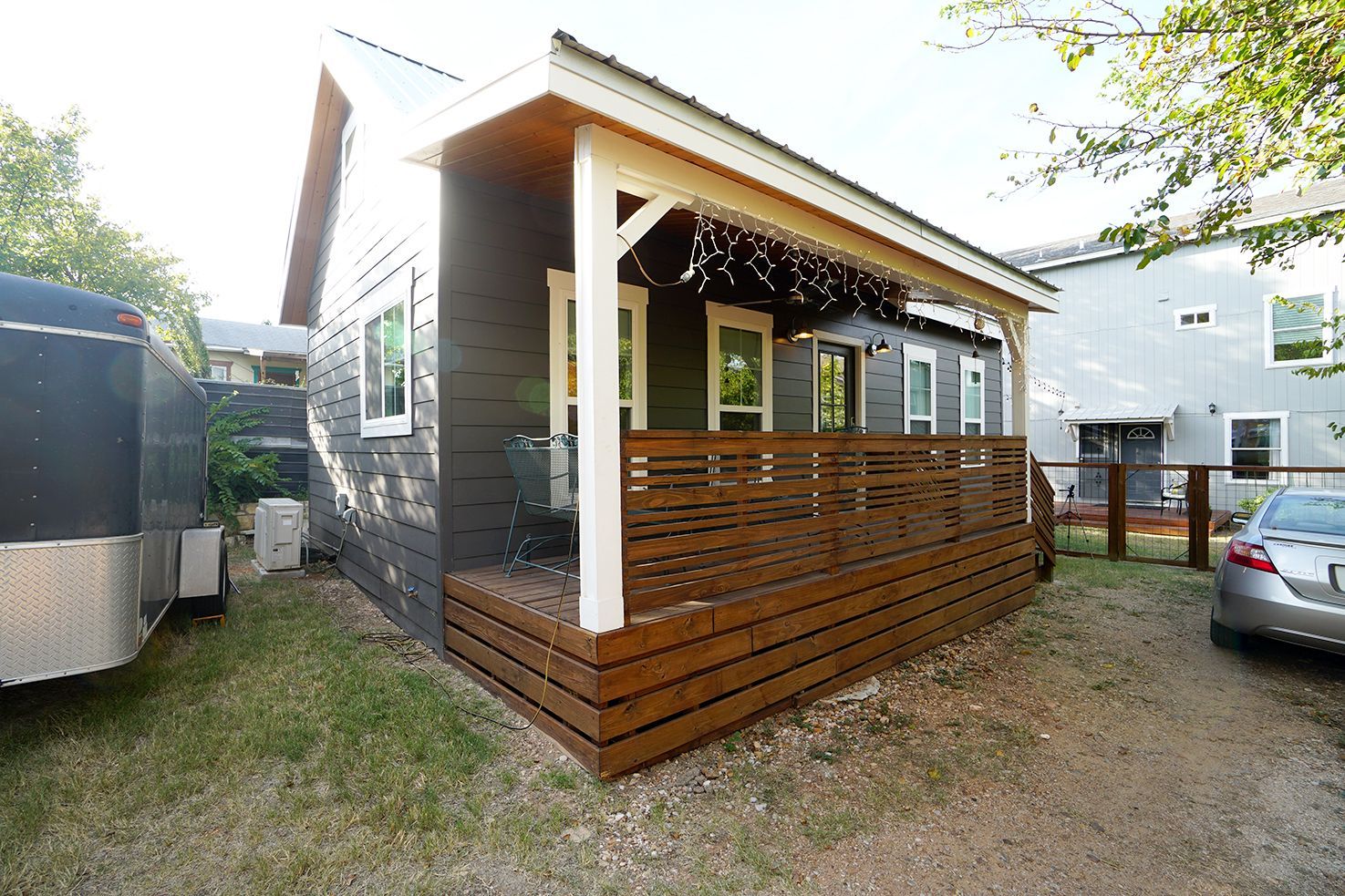 Small, dark-colored house with a wooden porch and railing, parked trailer next to it, car nearby.