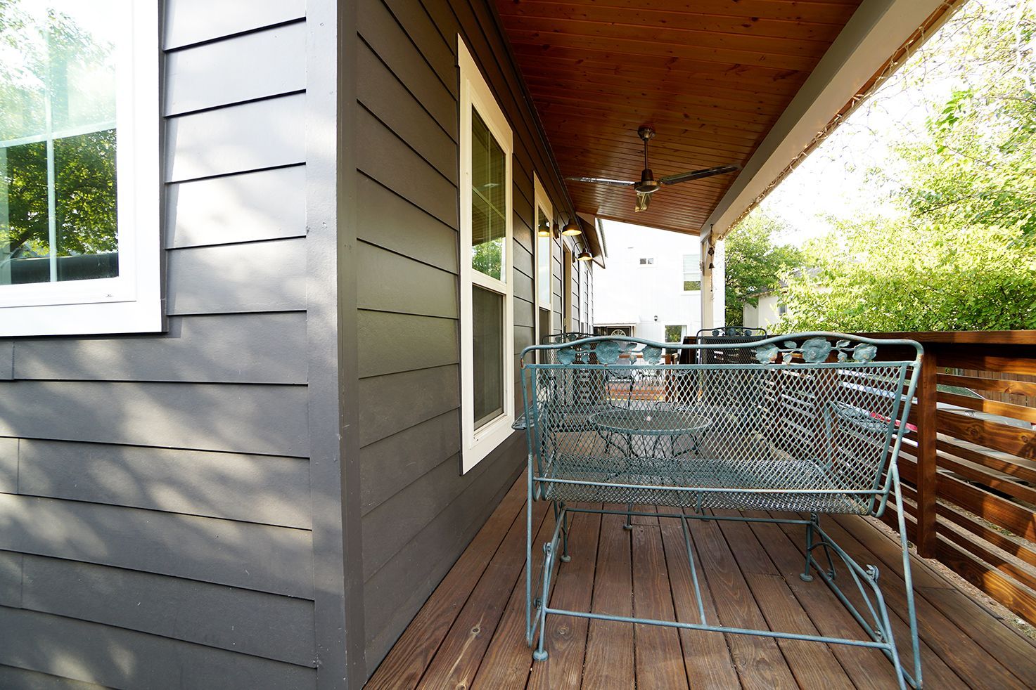 Exterior of a house with gray siding, a covered porch with a metal bench, and large windows.