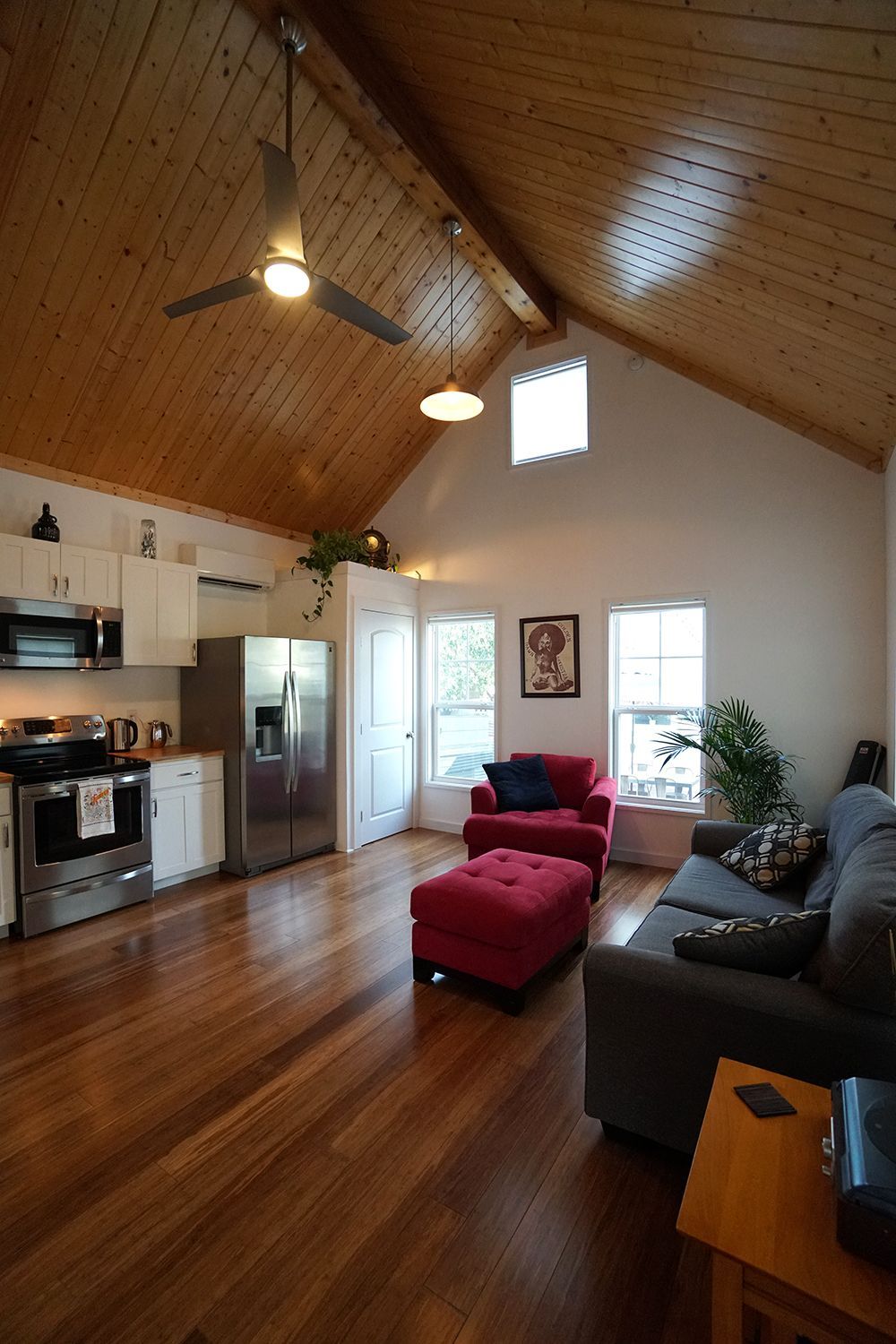 Living area with vaulted wood ceiling, open kitchen, stainless appliances, red ottoman and armchair, and gray sofa.