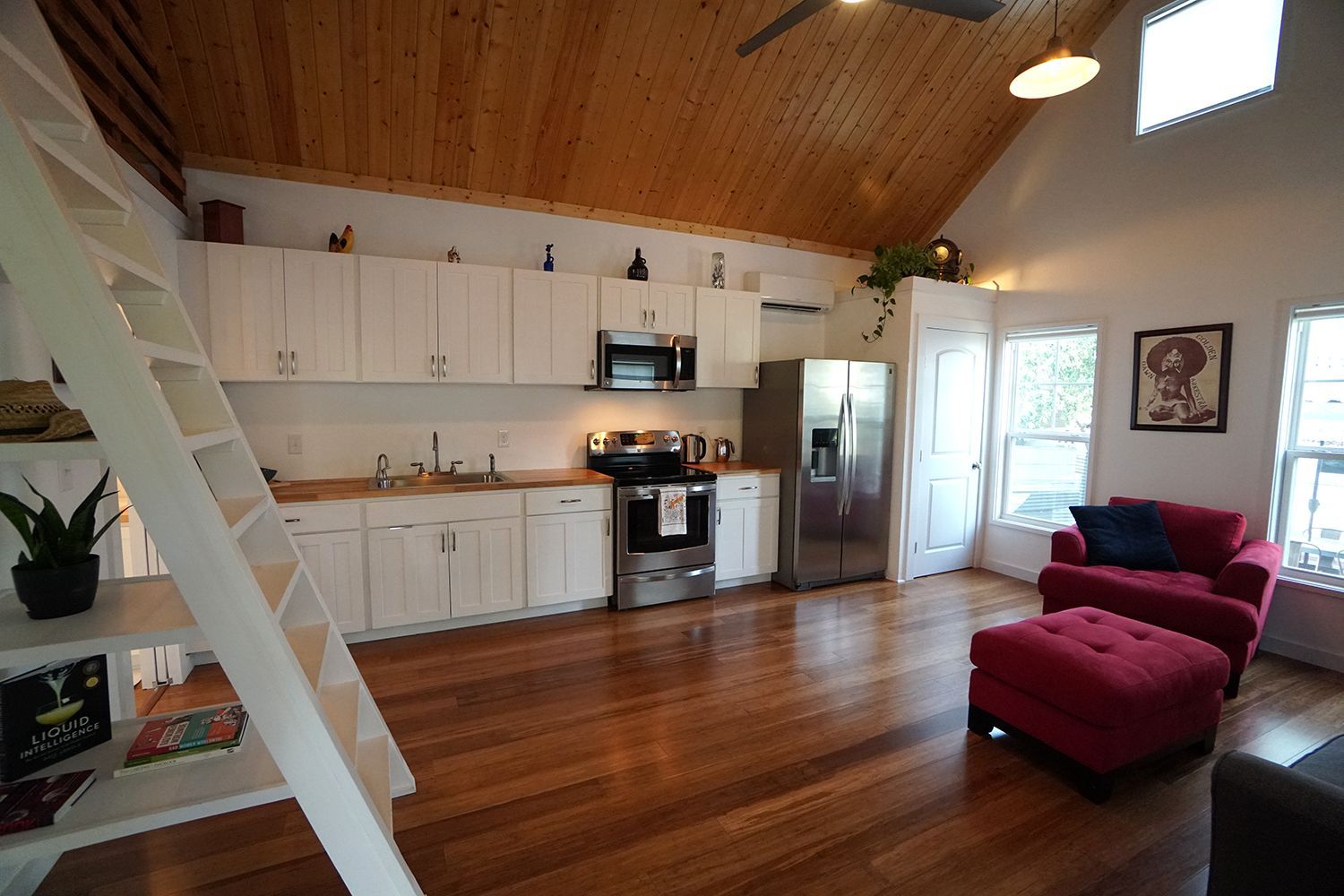Interior of a small apartment with kitchen, living area, and a ladder to a loft. Wooden floors and white cabinets.