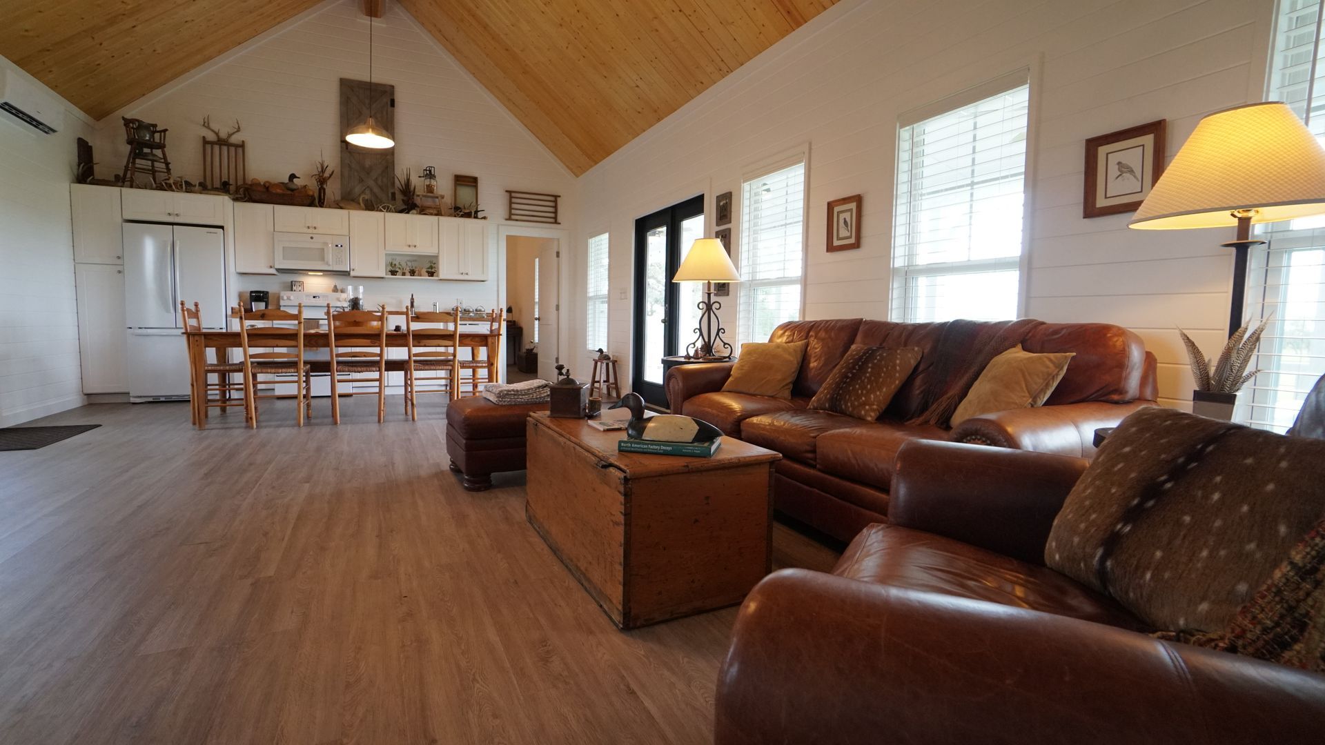Open-plan living space with leather furniture, wooden floor and high ceiling with kitchen in the background.