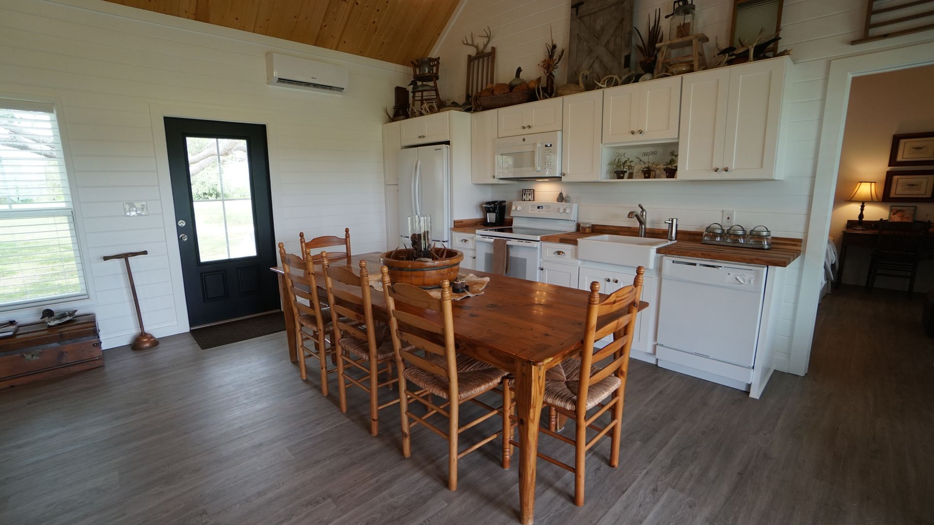 Kitchen with long wooden table and chairs, white cabinets, black door, and a glimpse into another room.