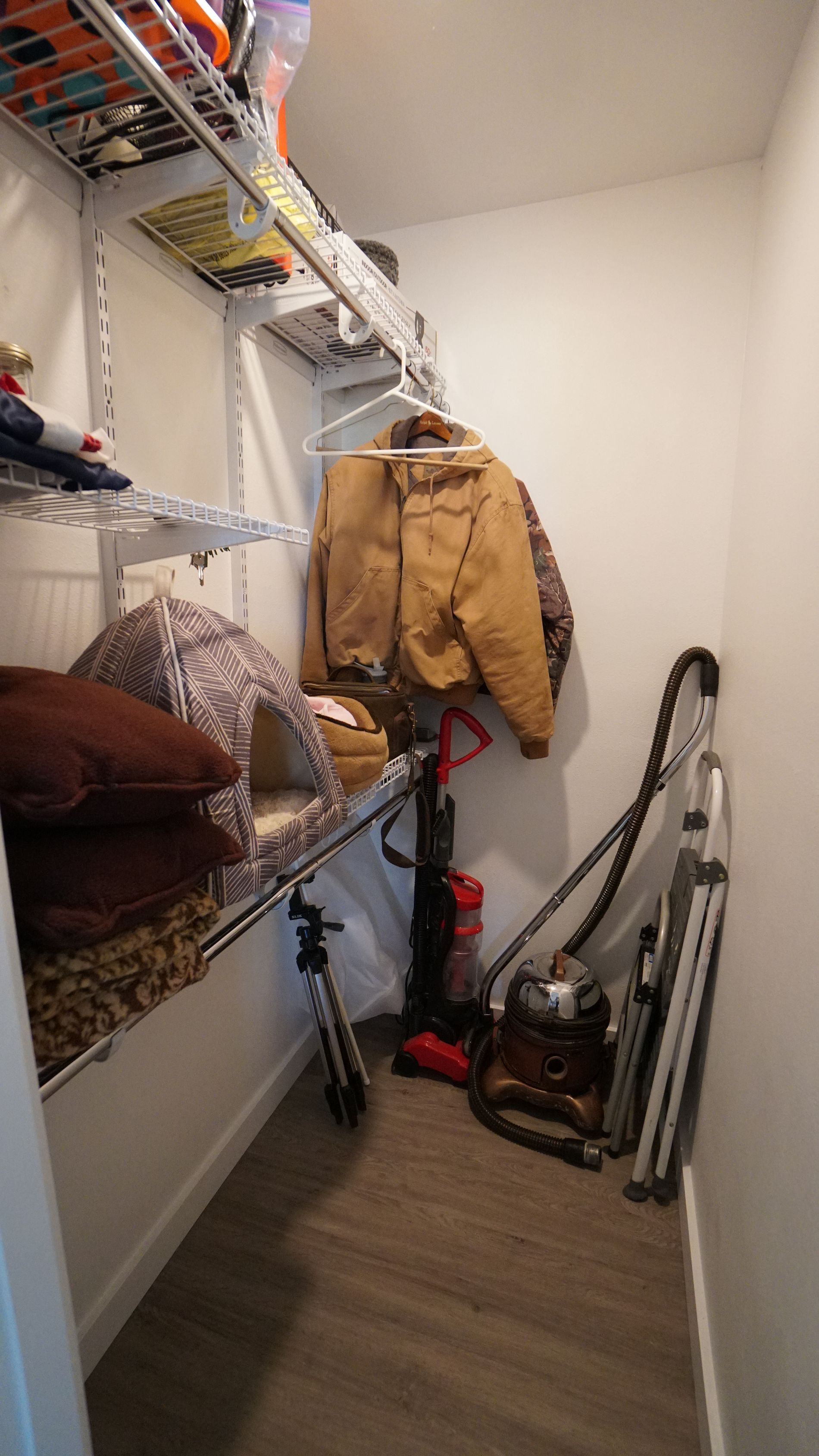 Small closet with wire shelving holding items. Jackets hang, vacuum cleaners and other objects on floor.