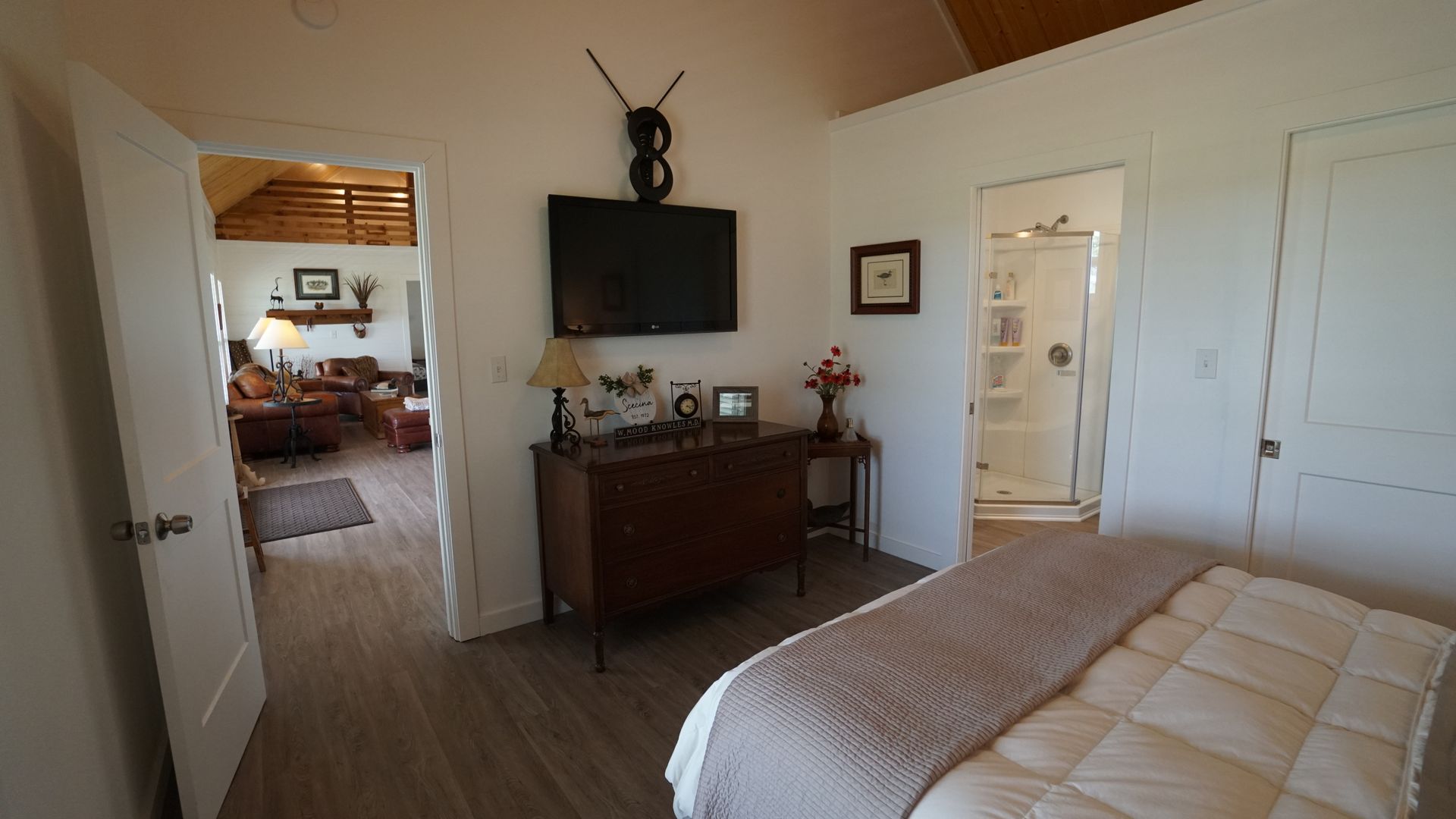 Bedroom with a view of a living room. TV above a dresser, a bed in the foreground, and a bathroom to the right.