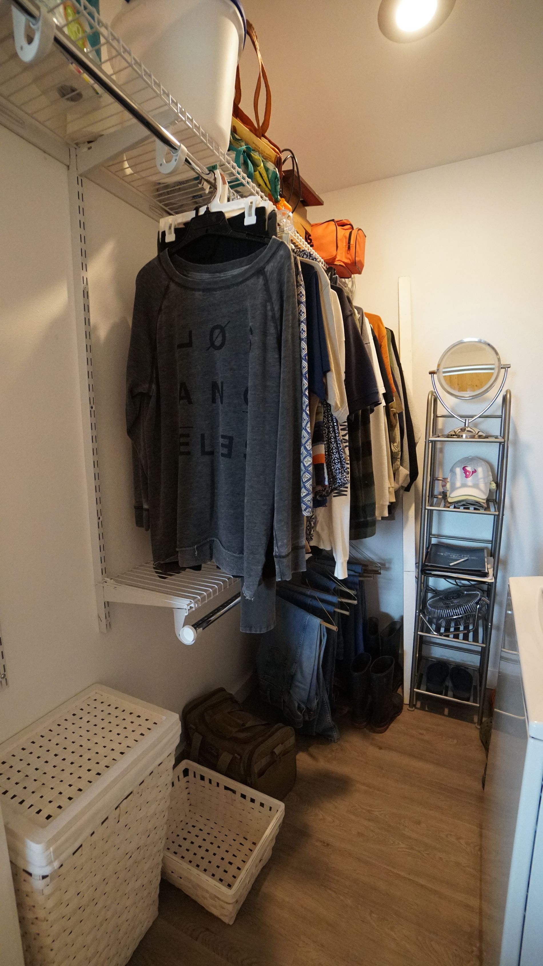 A small, cluttered closet with clothes on a rod, shelves, and baskets on the floor.