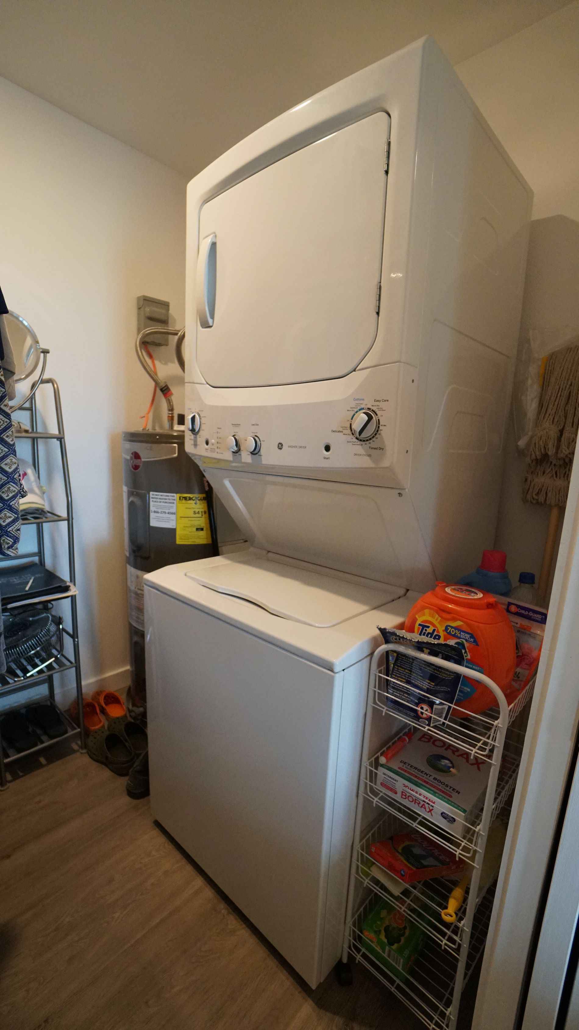 Stacked white washing machine and dryer in a laundry room, with shelving for detergent and other supplies.
