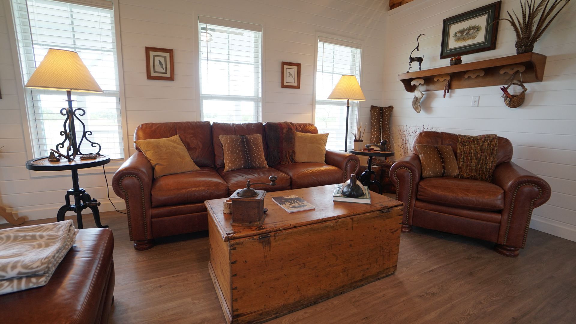 Cozy living room with brown leather sofa and armchair, wooden trunk coffee table, and lamps.