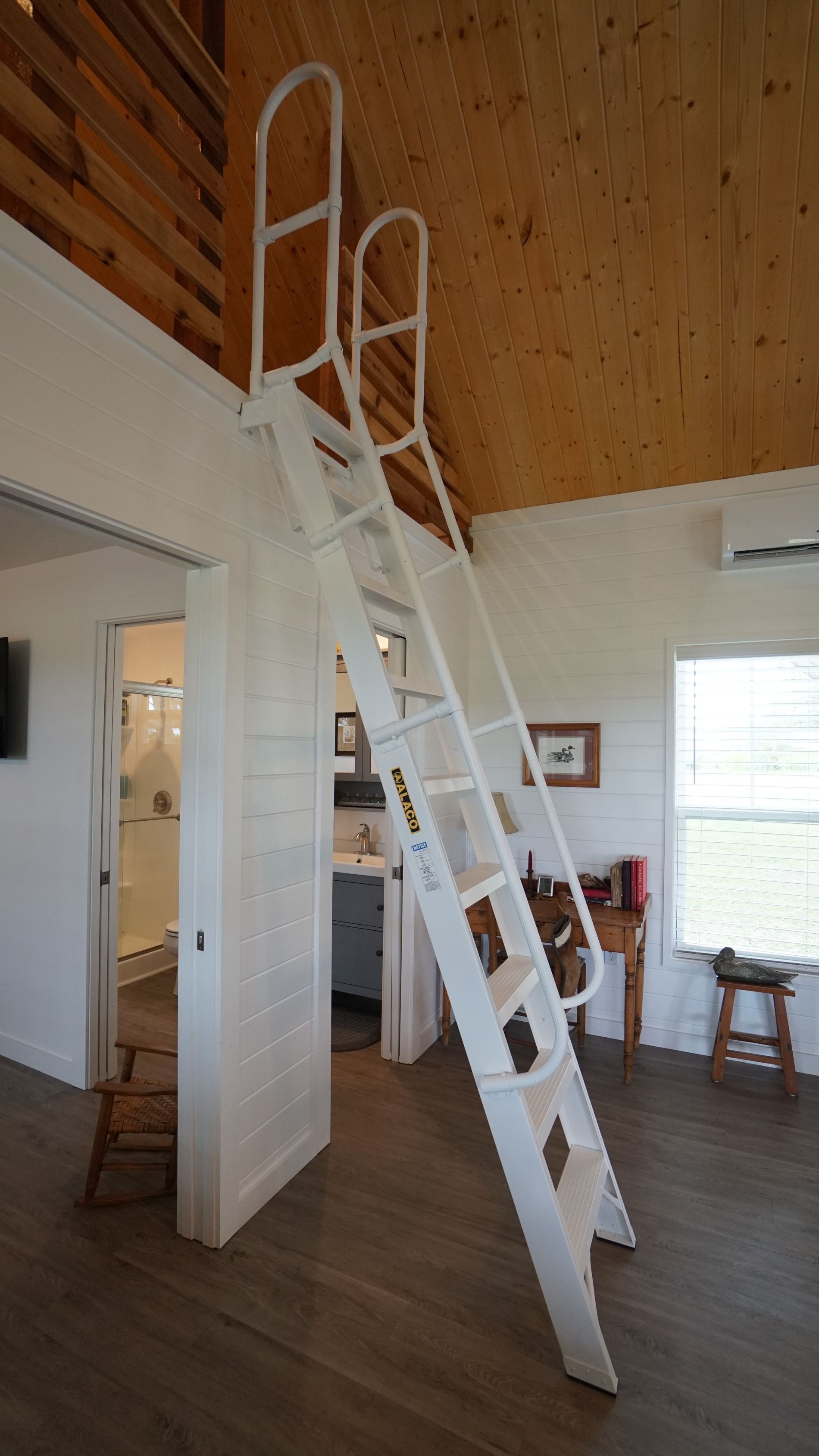 White angled ladder leading to a loft. Room with white walls, wood ceiling and a partially open bathroom door.