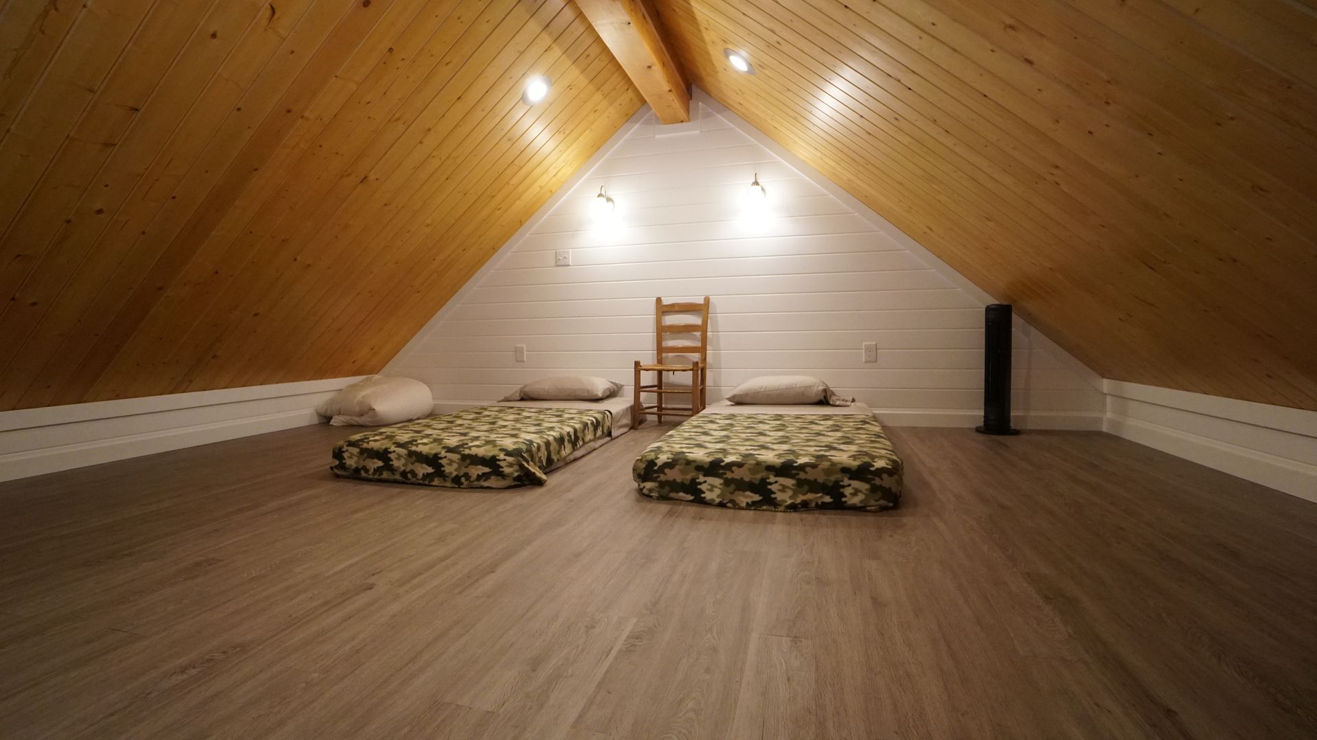 Attic bedroom with two mattresses on the floor, wooden ceiling and floor, white walls, and a ladder.