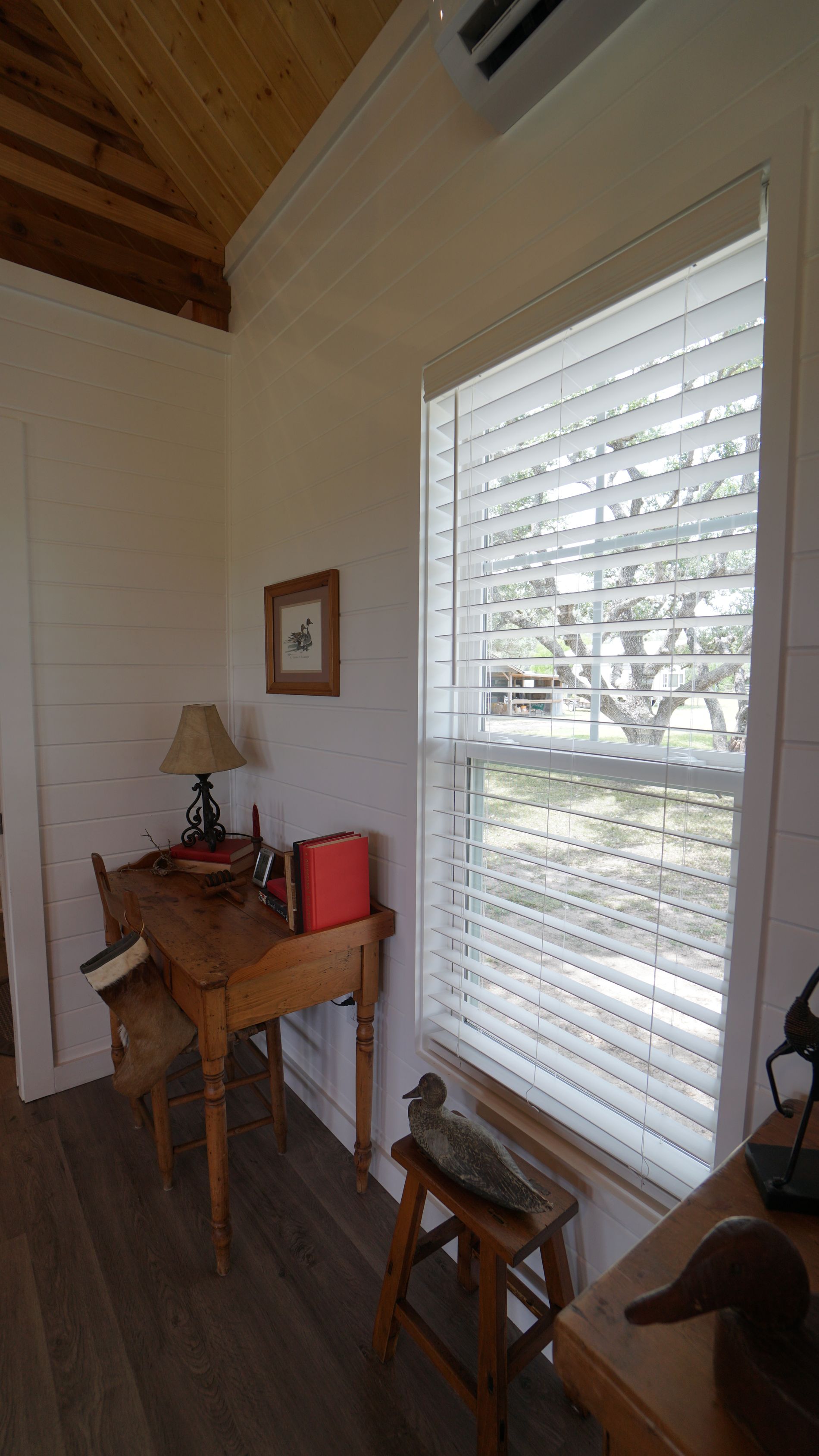 Small room with a window, desk, and duck decoy. White walls, wood floor, and sunlight.