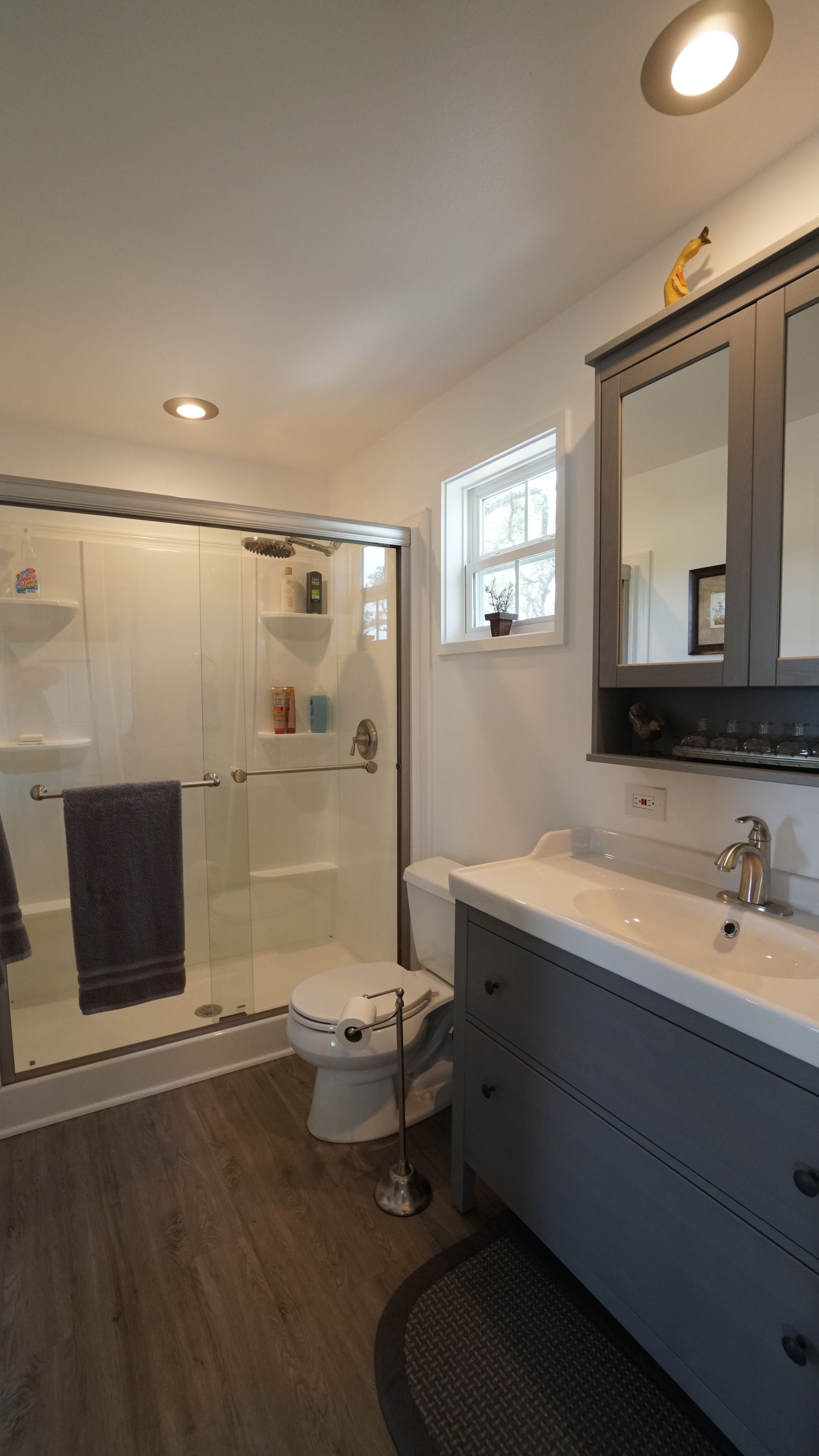 Bathroom with glass shower, gray vanity, toilet, and window, wood-look flooring.