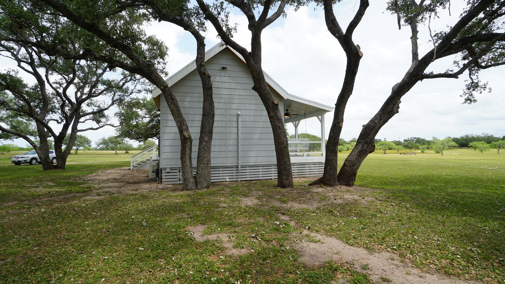 White wooden building shaded by trees in a grassy field, under an overcast sky.