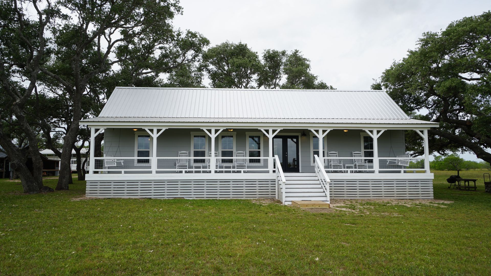 Gray house with white porch, metal roof, and ramp on a grassy lot with trees.