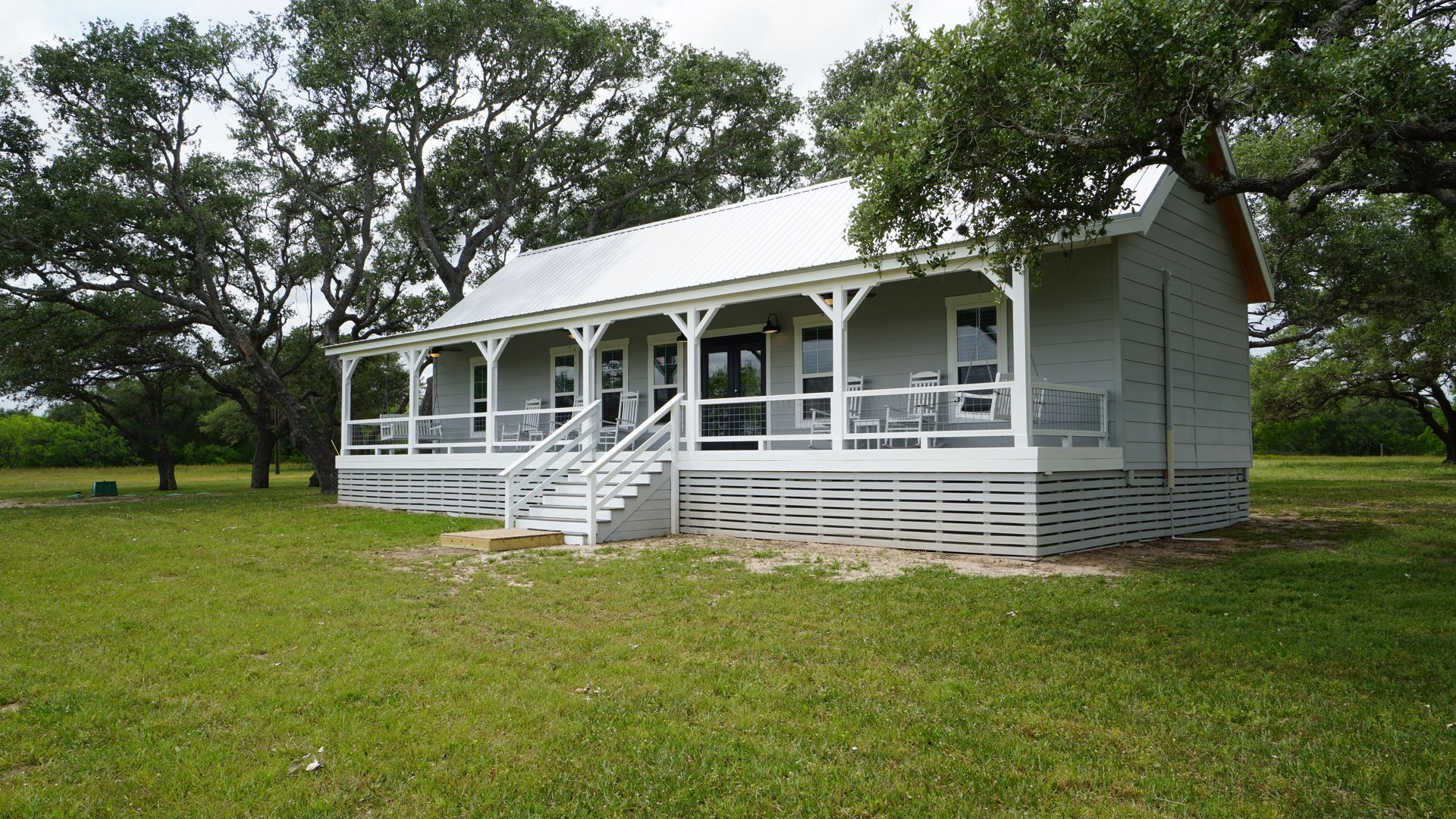 Gray cottage with white porch and metal roof in a grassy field, trees in background.