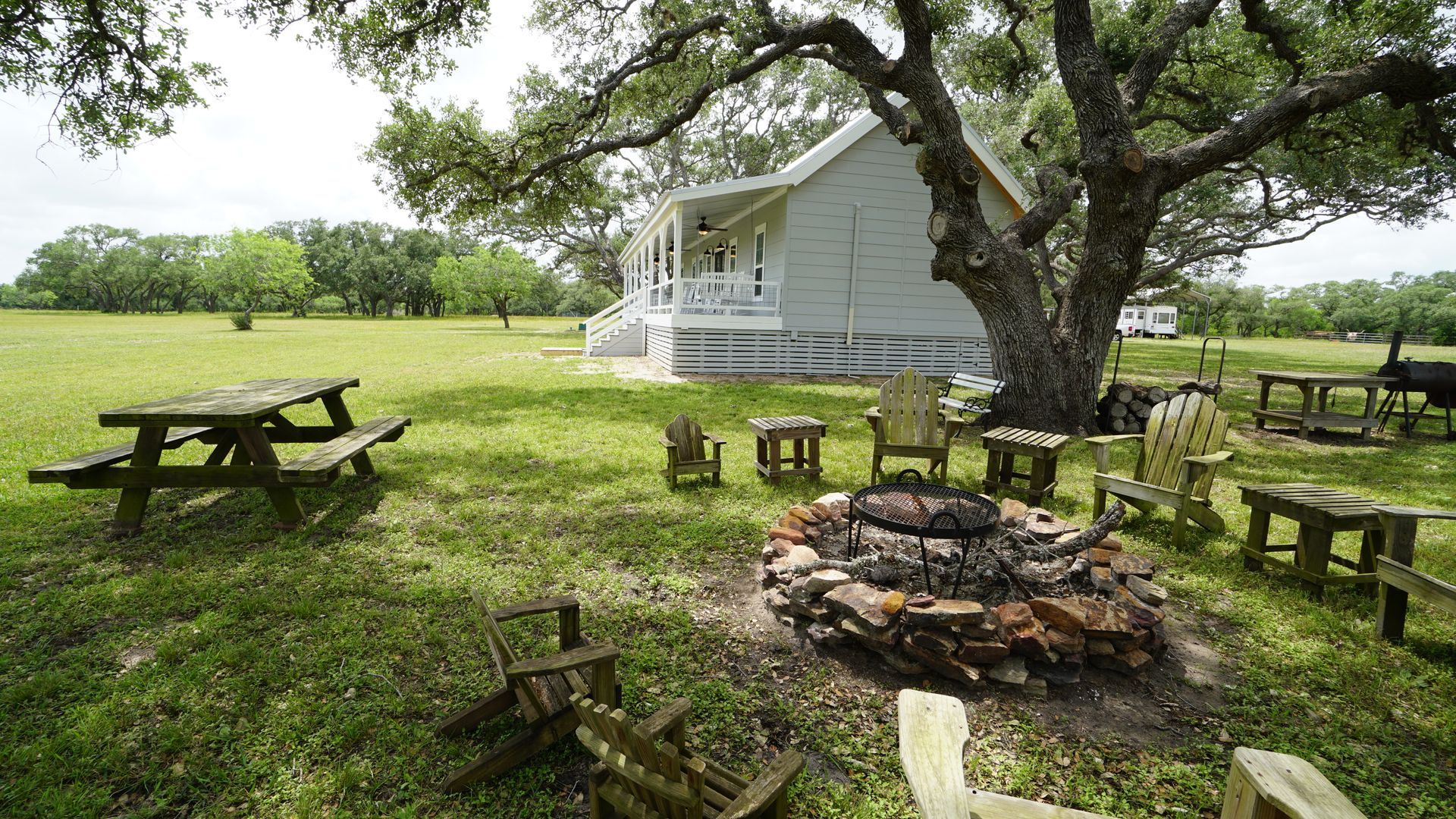 Lawn with a picnic table, chairs around a fire pit, and a white house under a large tree.