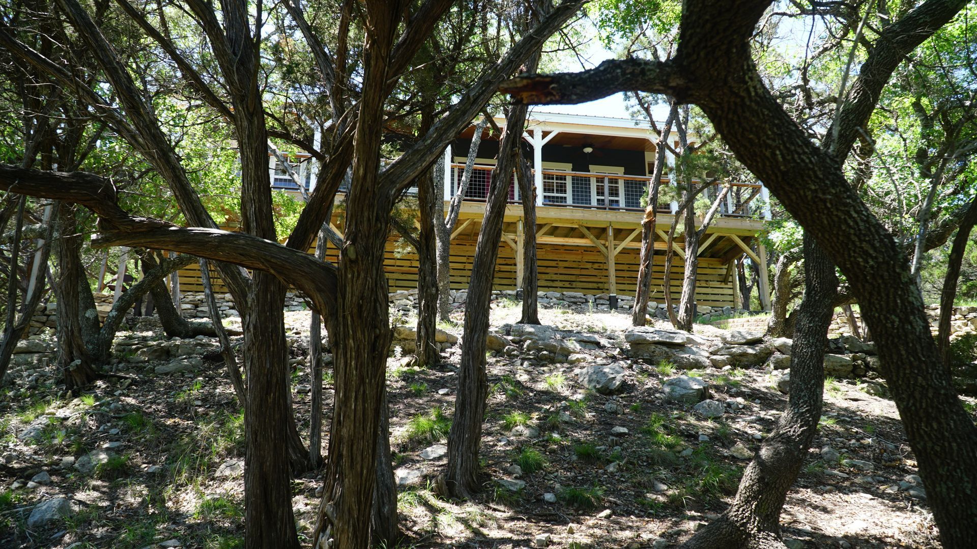 Cabin on stilts in a forest, framed by trees. Wooden deck, brown exterior, blue sky visible.