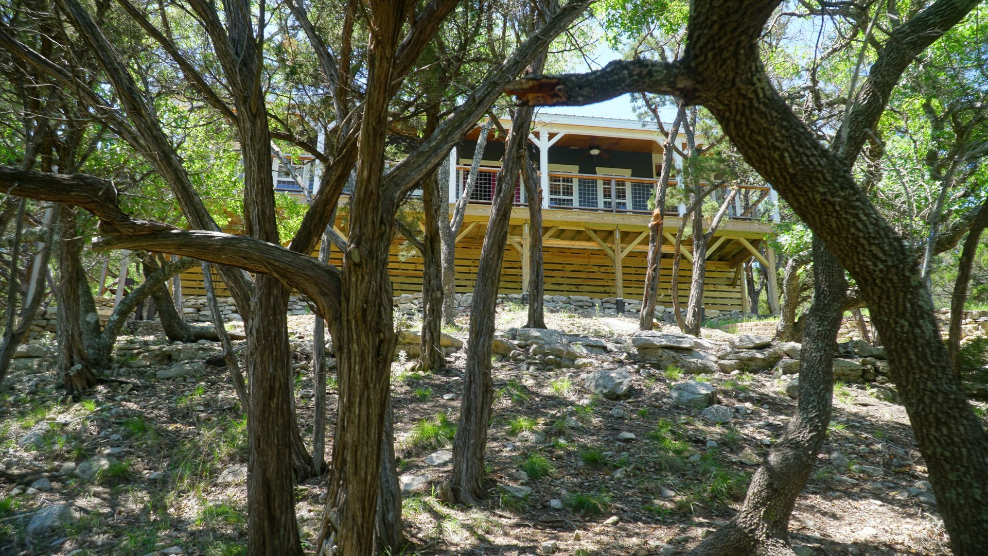 A wooden cabin with a large deck is nestled among trees on a rocky hill.