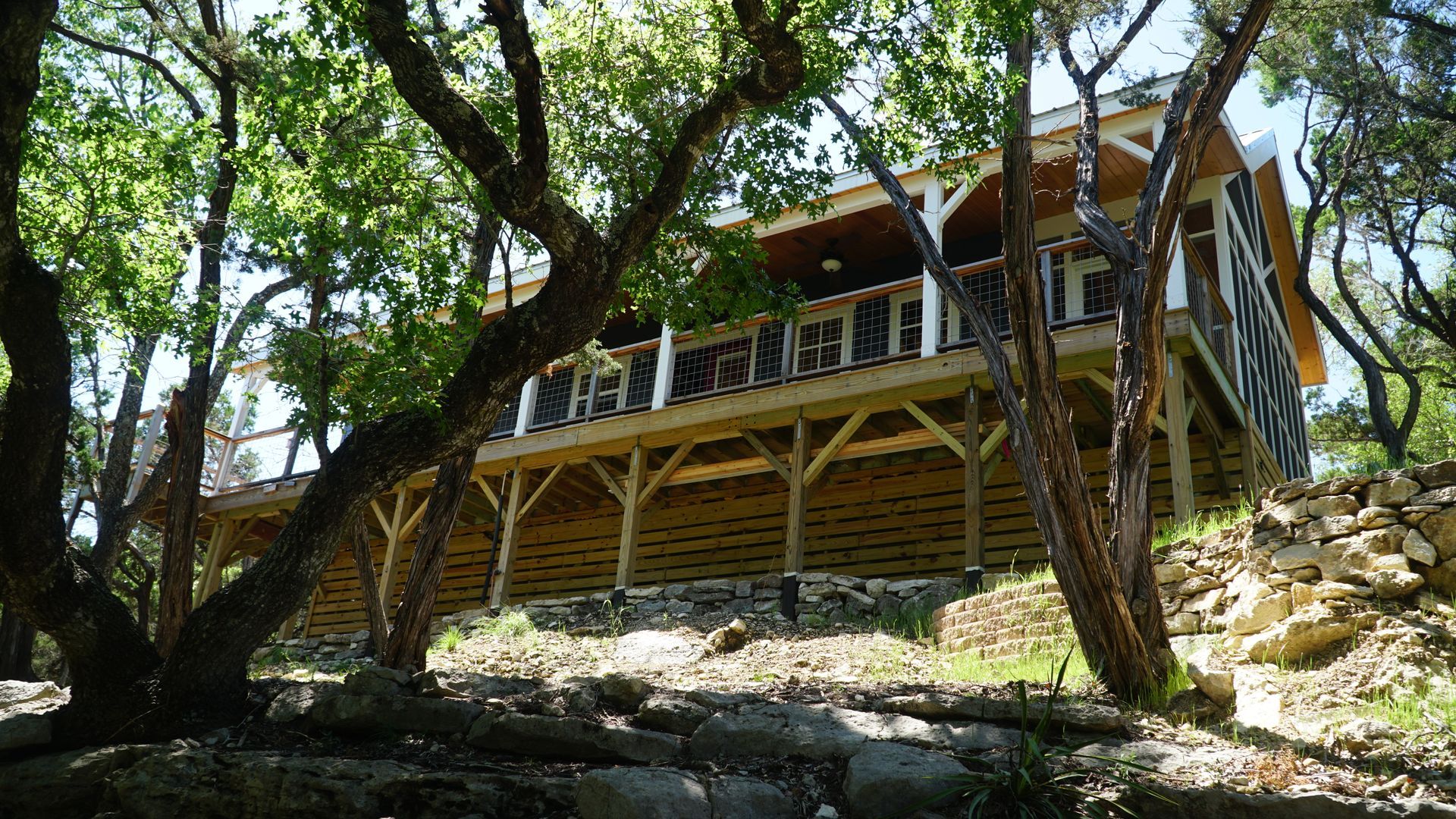 Wooden house on stilts in a forest setting, partially obscured by tree trunks and branches.