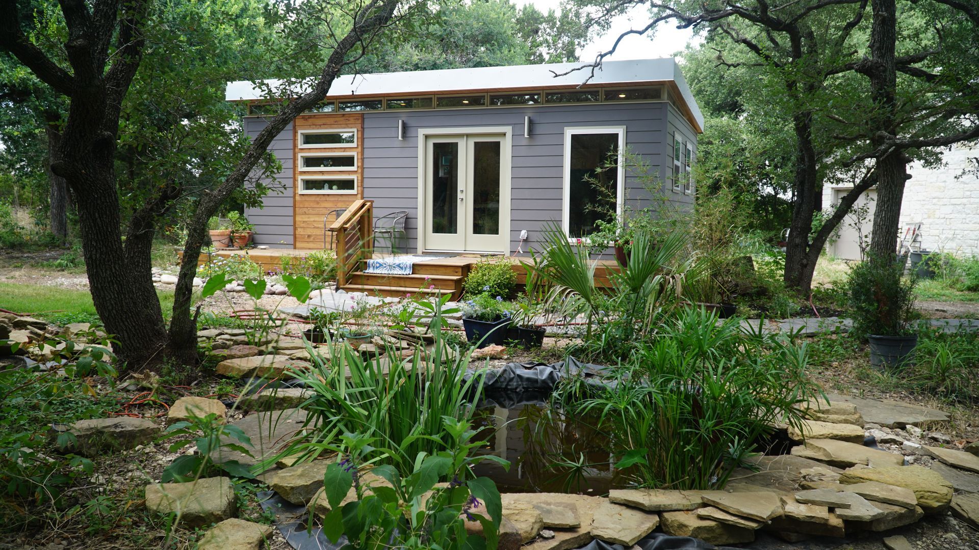 Small gray cabin with double doors, windows, and wooden accents, surrounded by a garden with a small pond.
