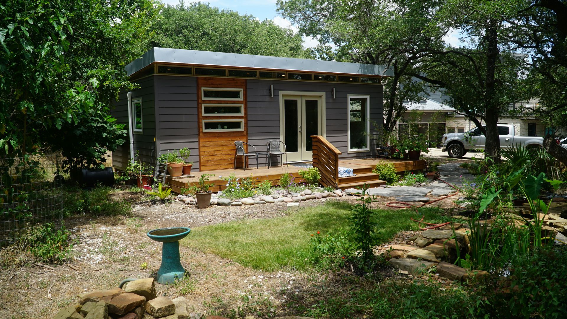 Gray tiny home with wooden deck, surrounded by greenery.