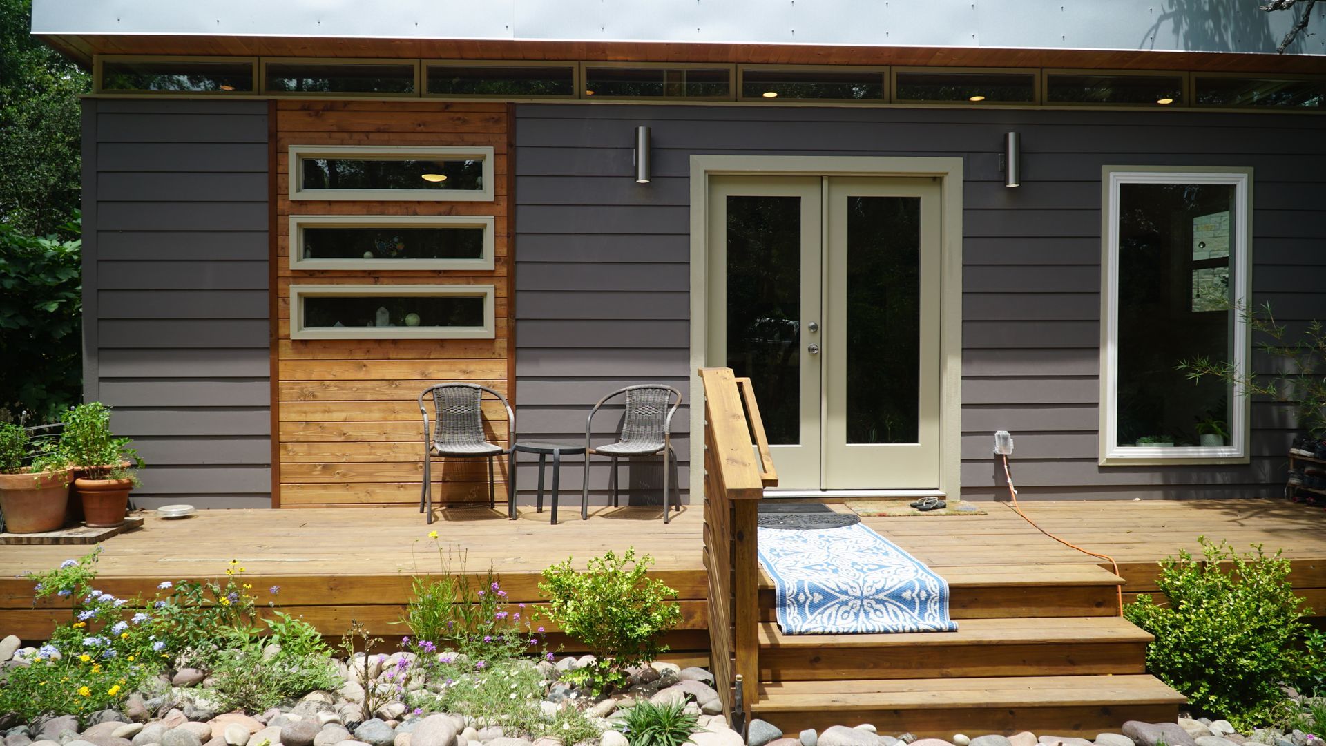 Modern tiny house with wooden deck, gray siding, and double doors; chairs and rug on deck.