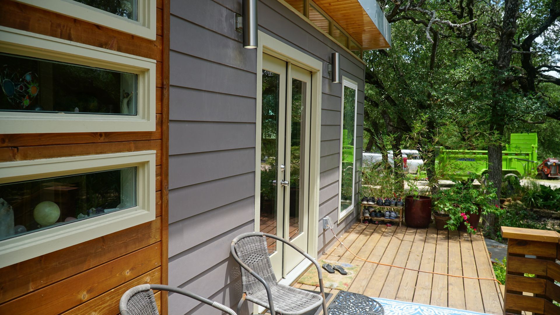 Exterior view of a small home with grey siding, windows, and a wooden deck with chairs.