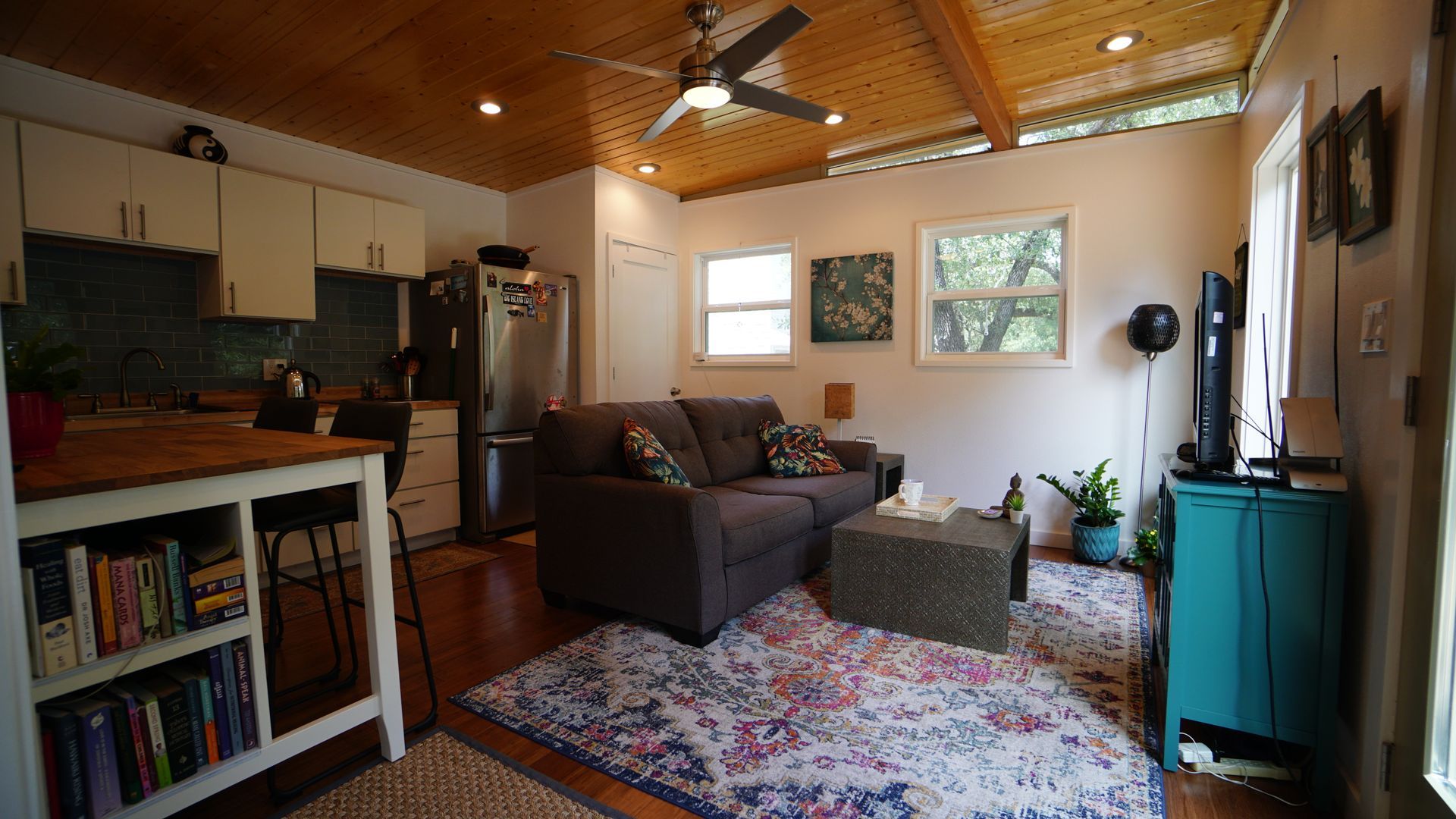 Cozy living space with kitchen, couch, and teal cabinet. Wooden ceiling, rug, and natural light.