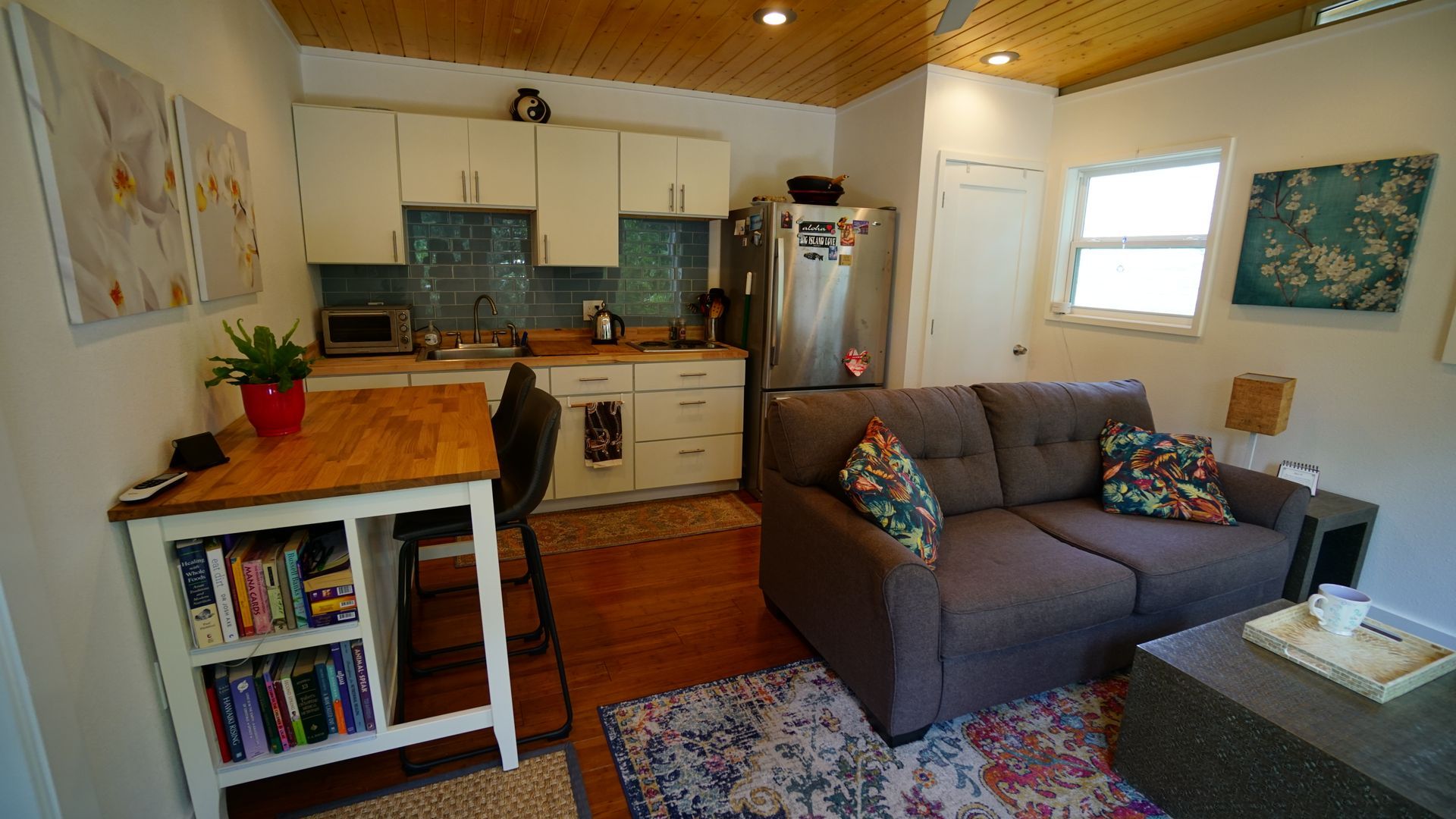 Small, cozy living space with kitchen and seating area. Wooden table, couch, cabinets, refrigerator, and decorative elements.