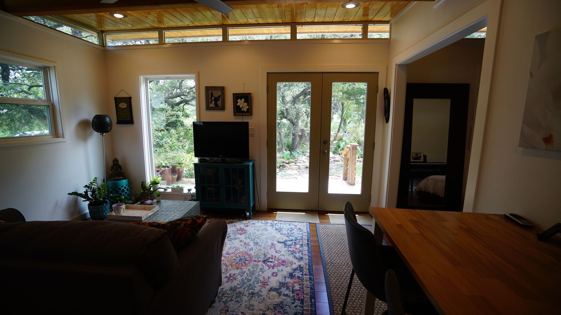 Cozy living room with large windows overlooking trees. Includes couch, TV, rug, and dining table.
