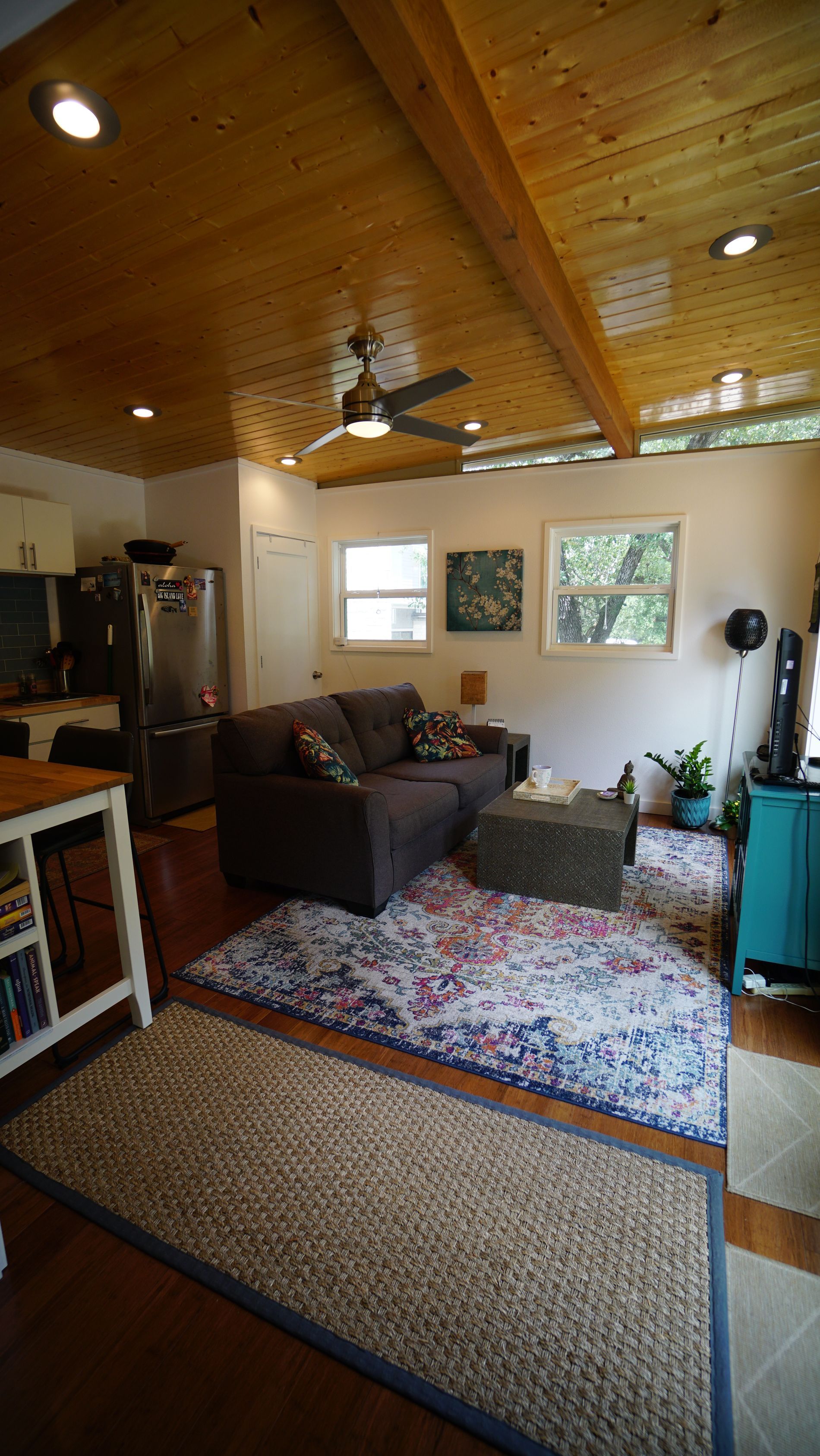 Living room with wooden ceiling, brown sofa, area rugs, and windows.