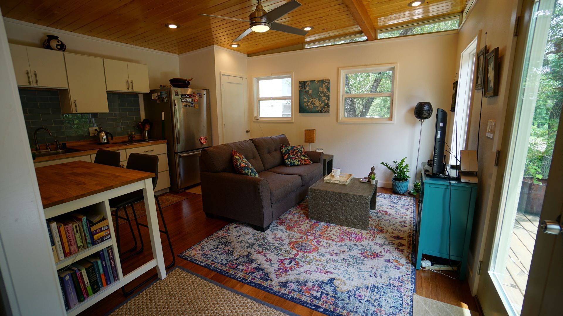 Cozy living space with kitchen, sofa, and teal storage cabinet. Light streams in from open door and windows.