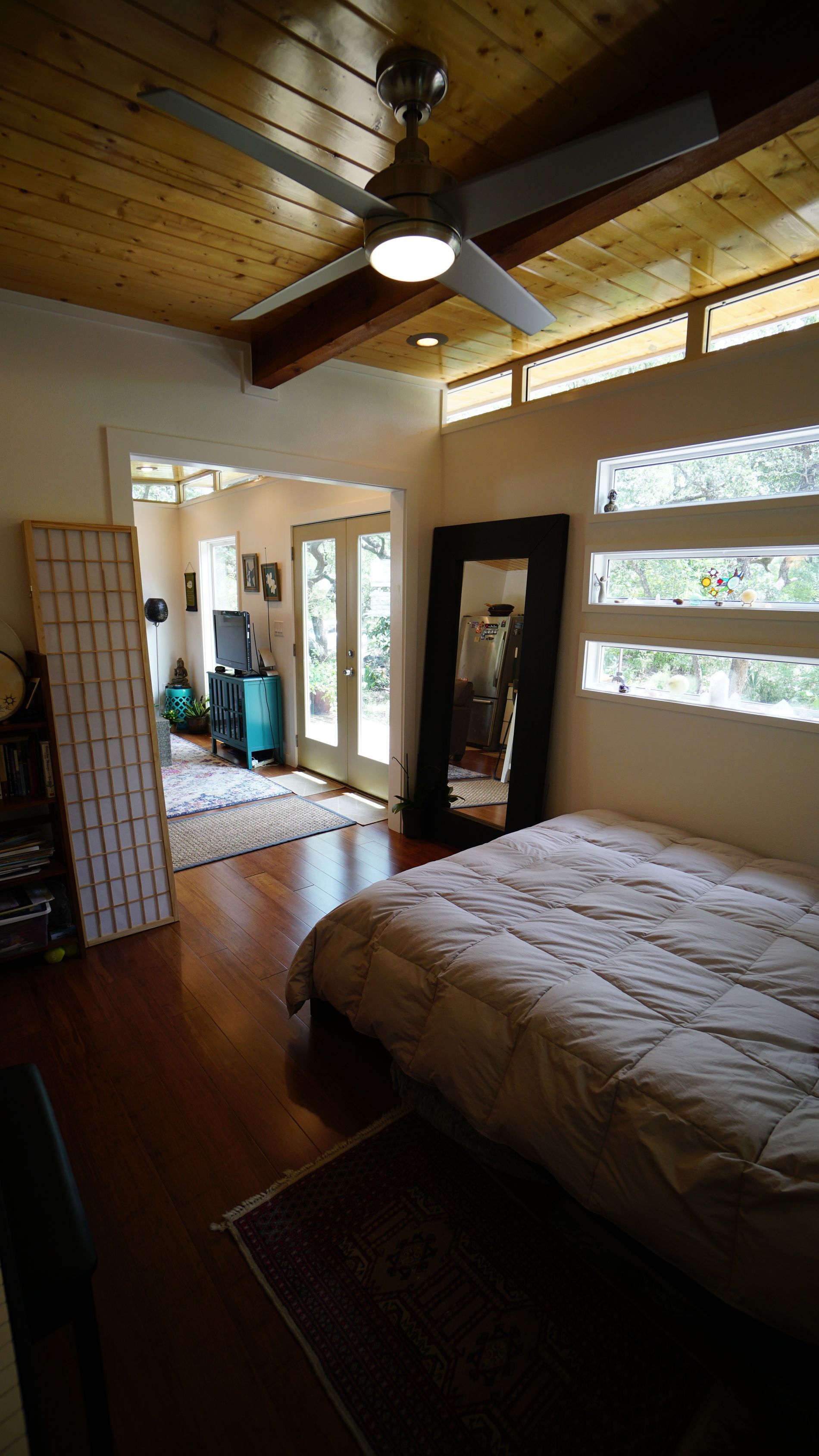 Bedroom with wooden floor, bed, full-length mirror, door leading to another room. Ceiling fan and windows visible.