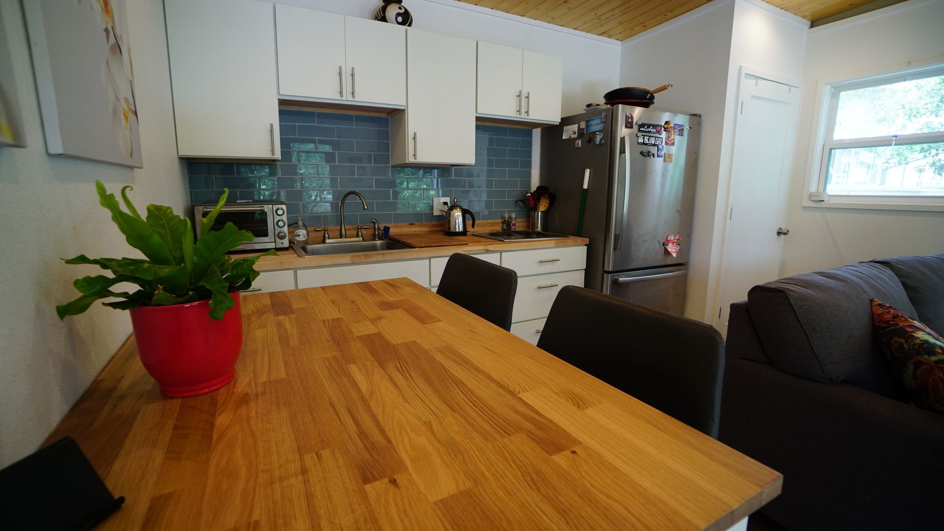 Small kitchen with wooden table, white cabinets, stainless steel fridge, and plant in red pot.