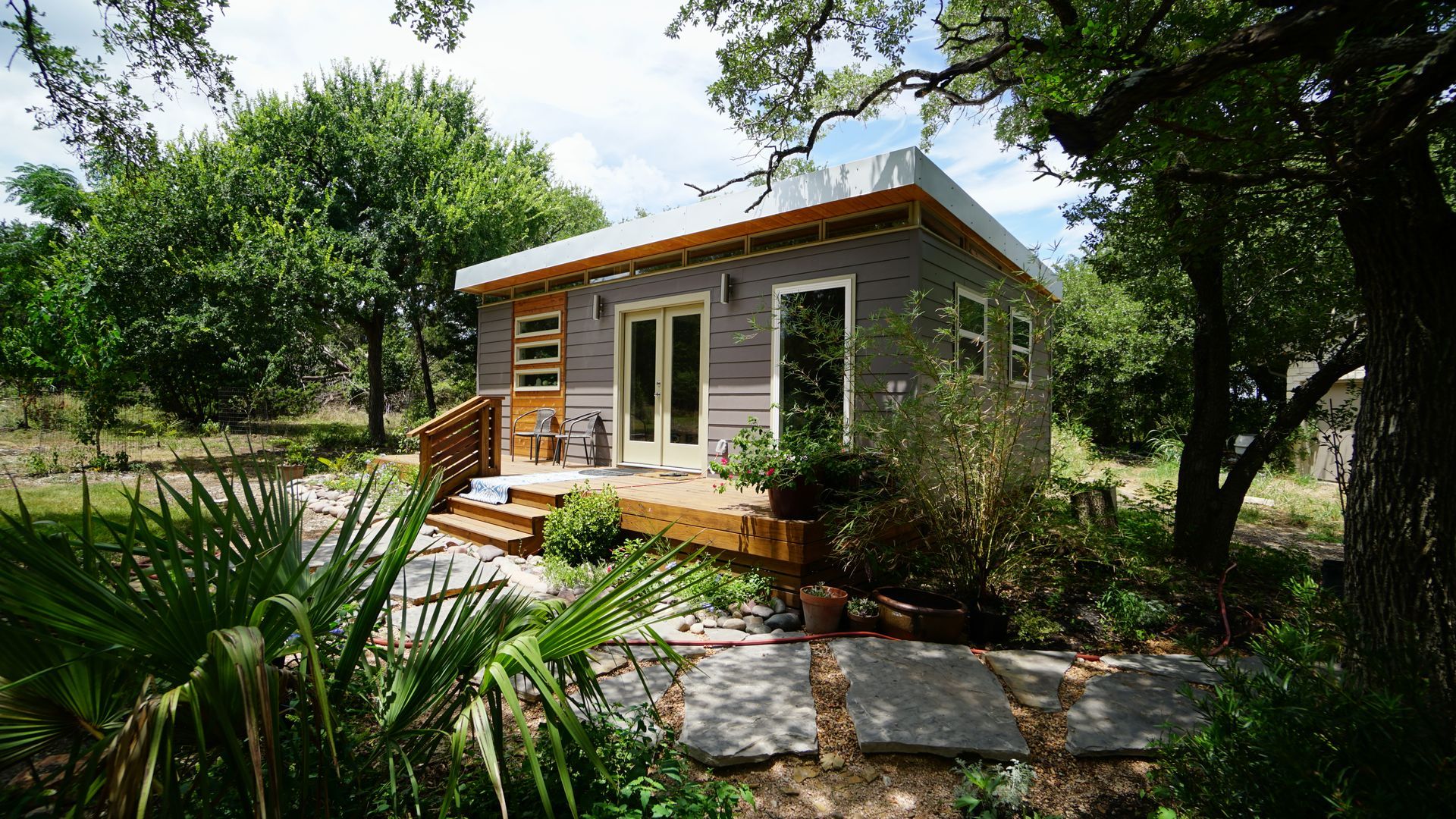 Small, modern cabin with a wooden deck, surrounded by trees and greenery.