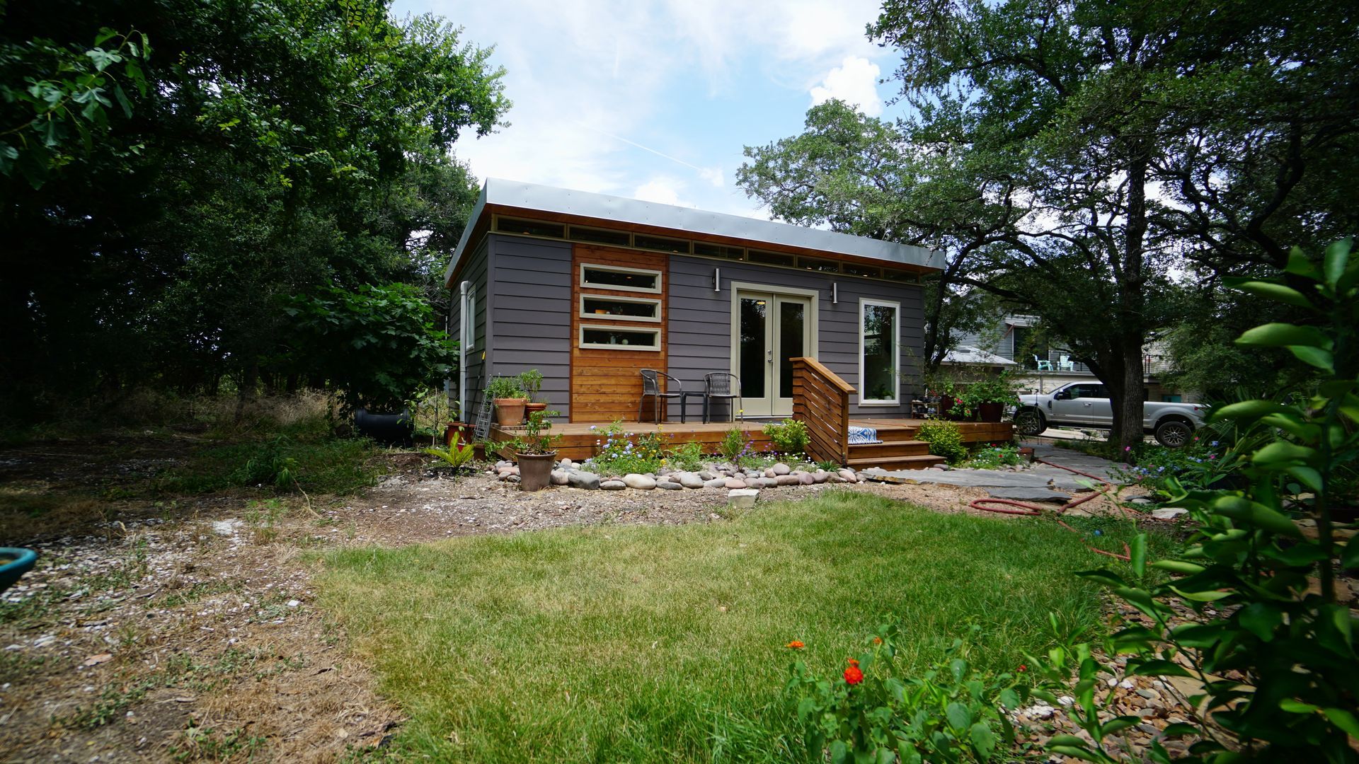 Small gray house with wooden accents and a front porch, surrounded by trees and a lawn.