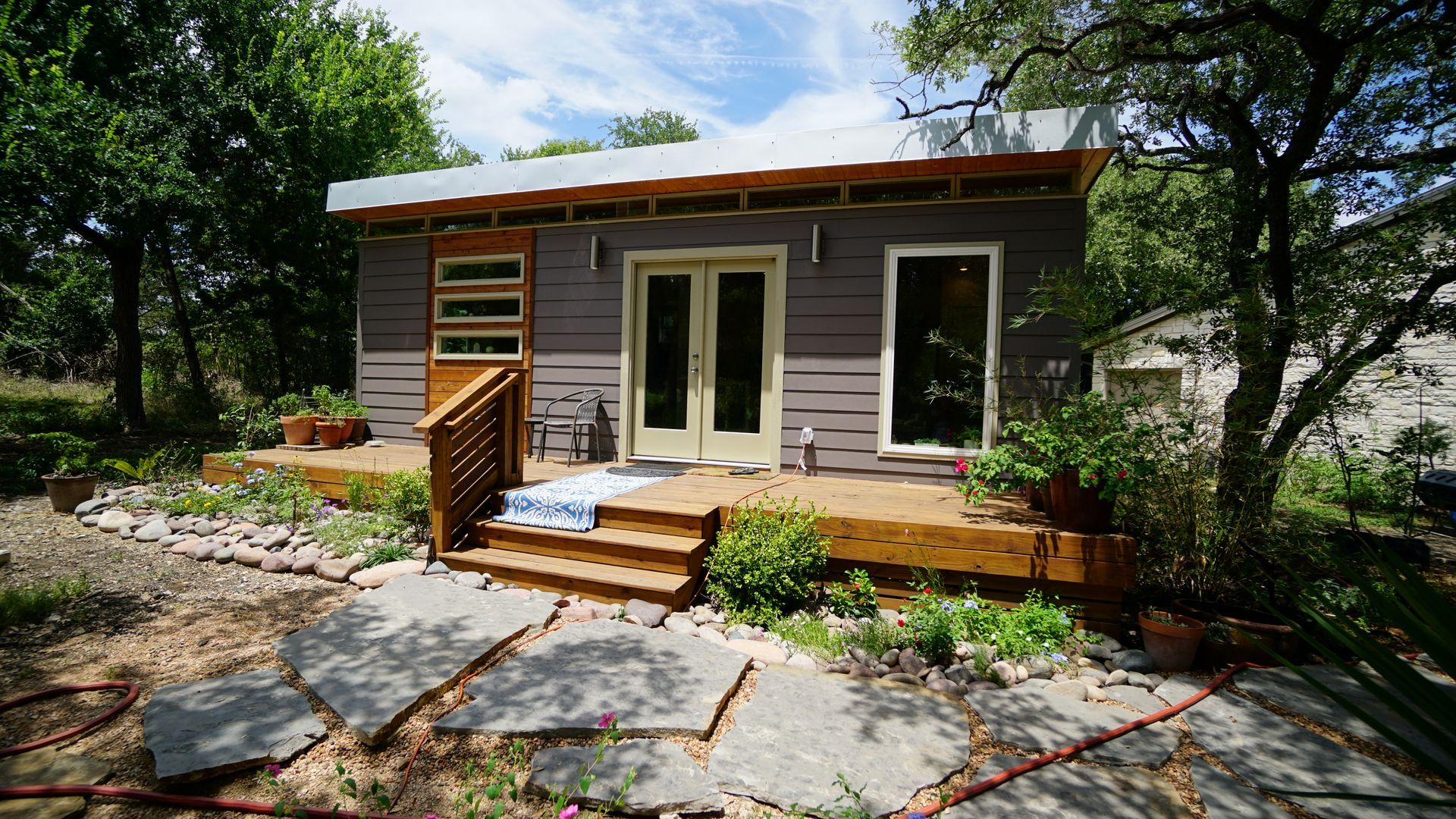 Small gray and wood-trimmed house with a wooden deck and stone path in a green, natural setting.