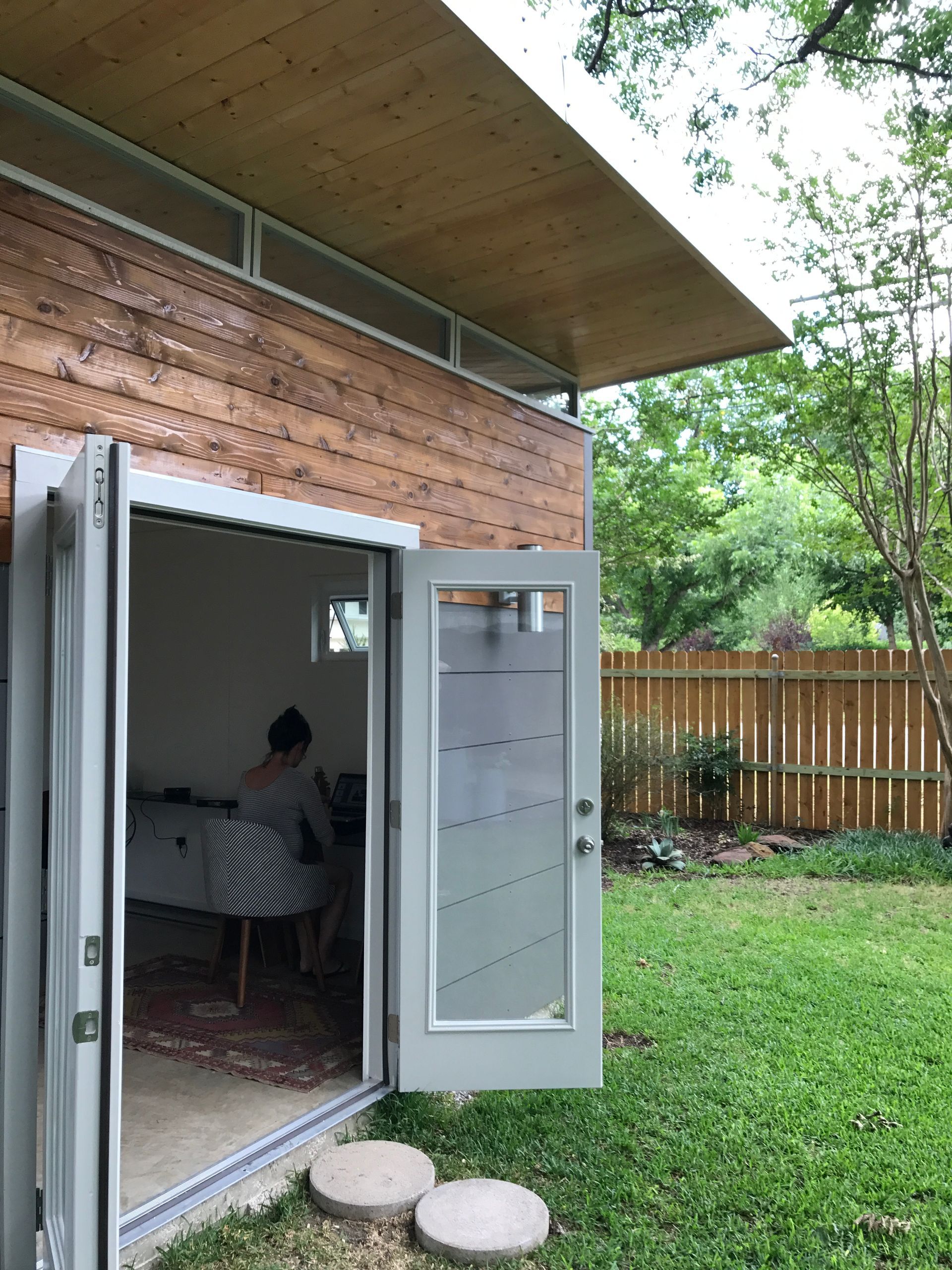 Modern backyard office with open doors, person working inside, wooden siding, and green lawn.