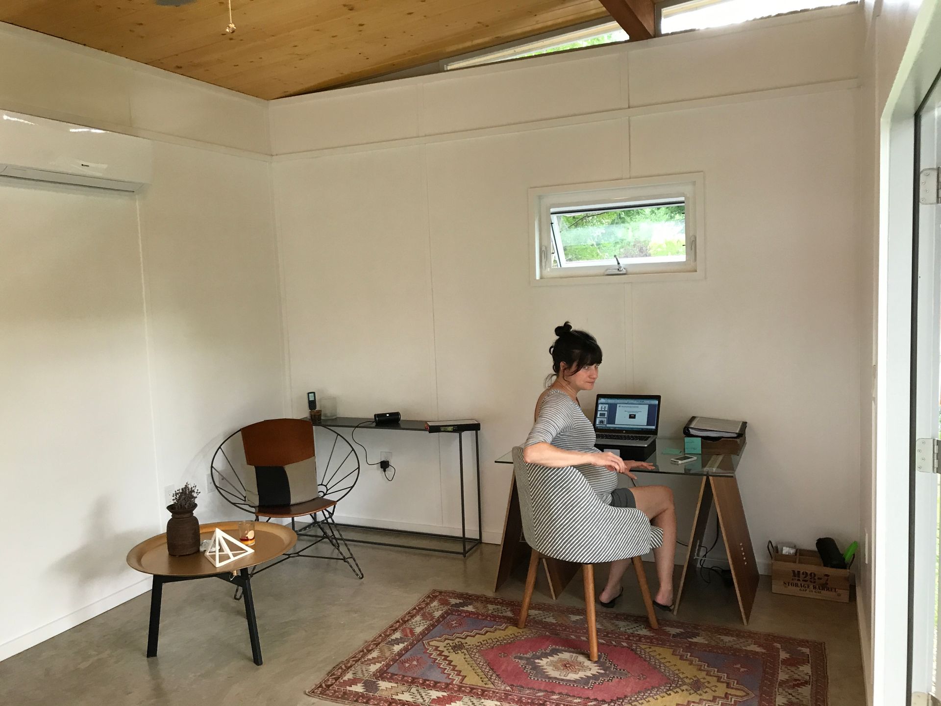 Woman working on laptop at a desk in a small, well-lit room with a small window, chair, and rug.