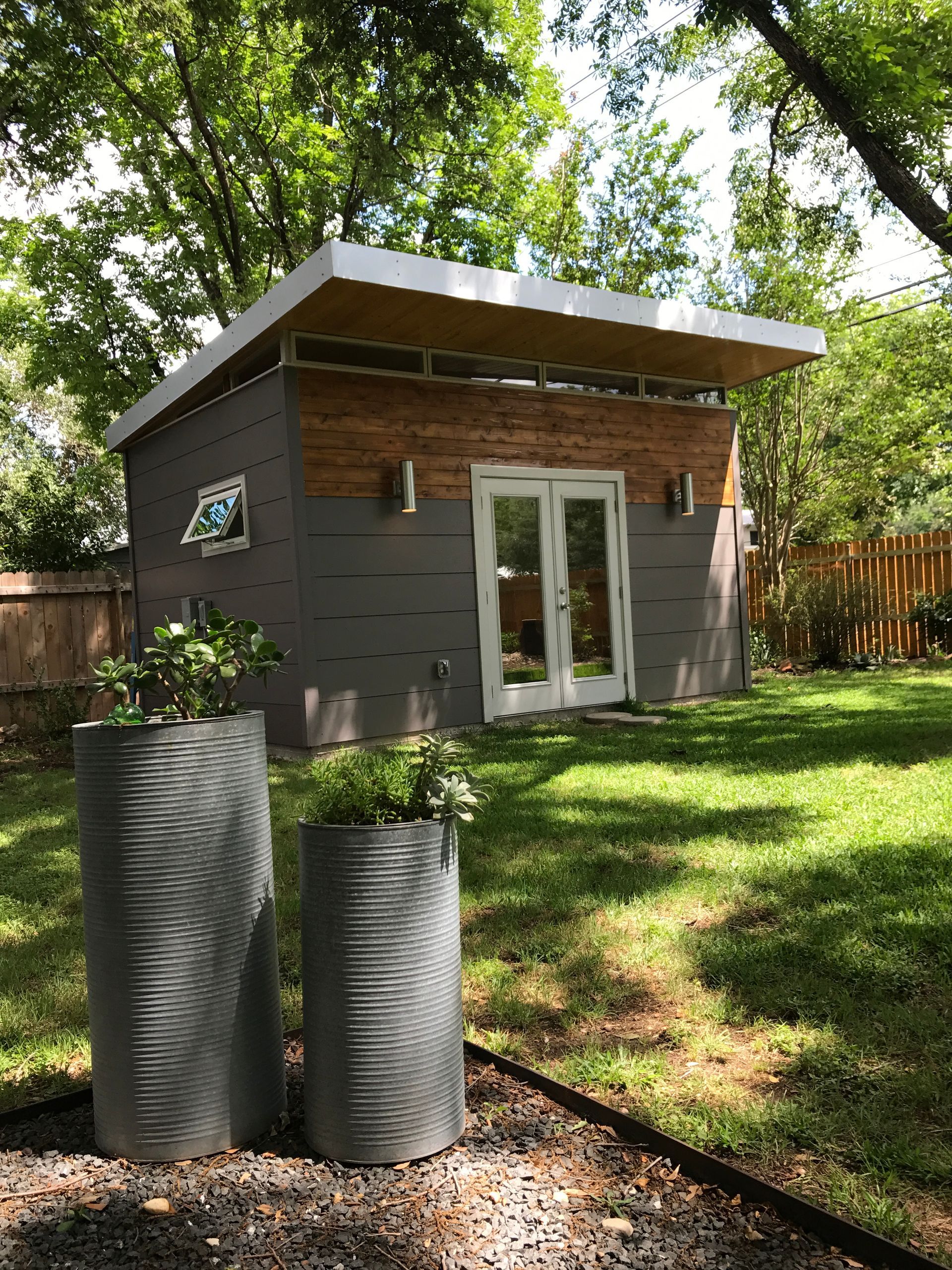 Modern backyard studio with gray siding, wood accent, and double doors, flanked by planters.