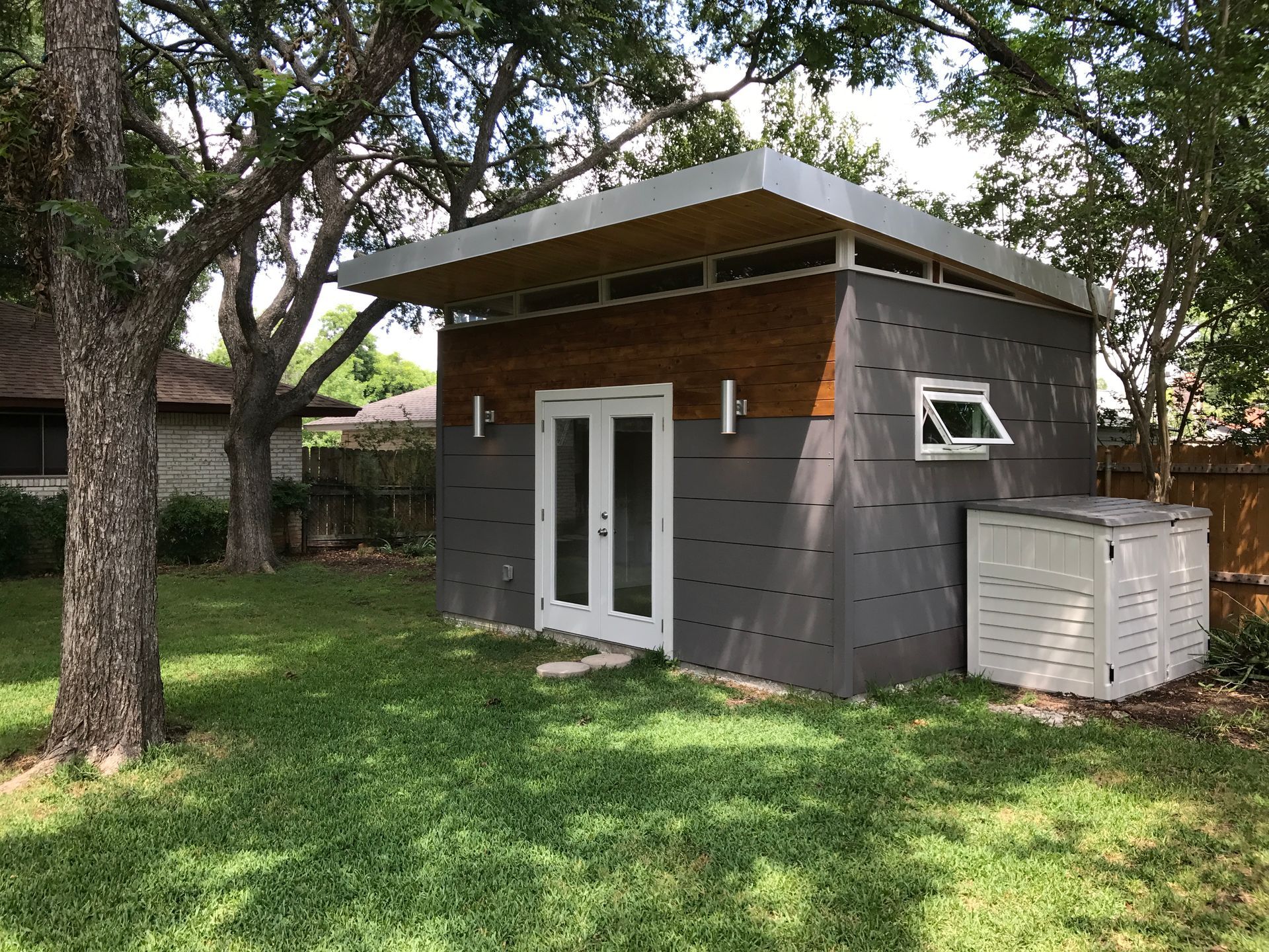 Modern gray and brown shed with white double doors, small window, and air conditioning unit in a grassy yard.
