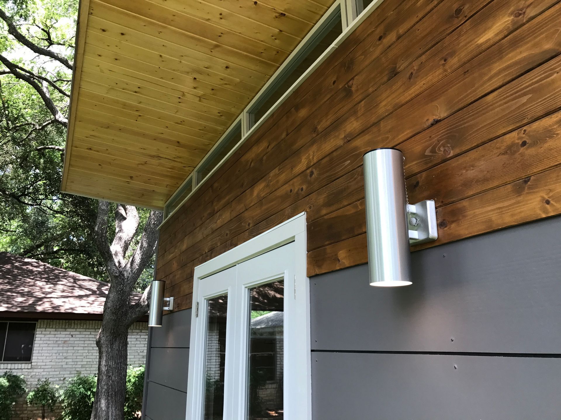 Exterior view of a building with wooden siding, silver lights, and white doors. Grey and tan colors.