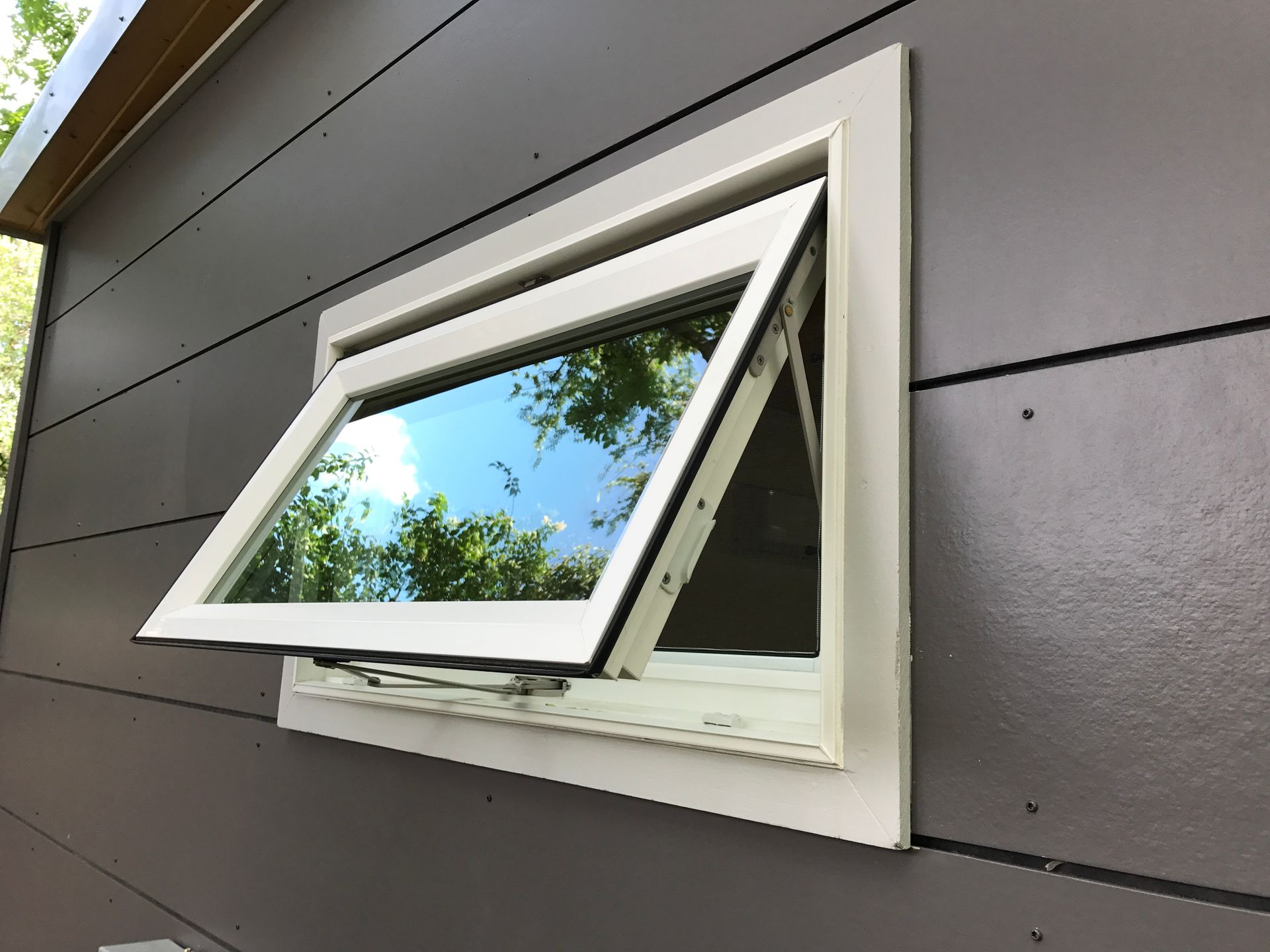 Open, white-framed window on a gray building reflecting sky and trees.