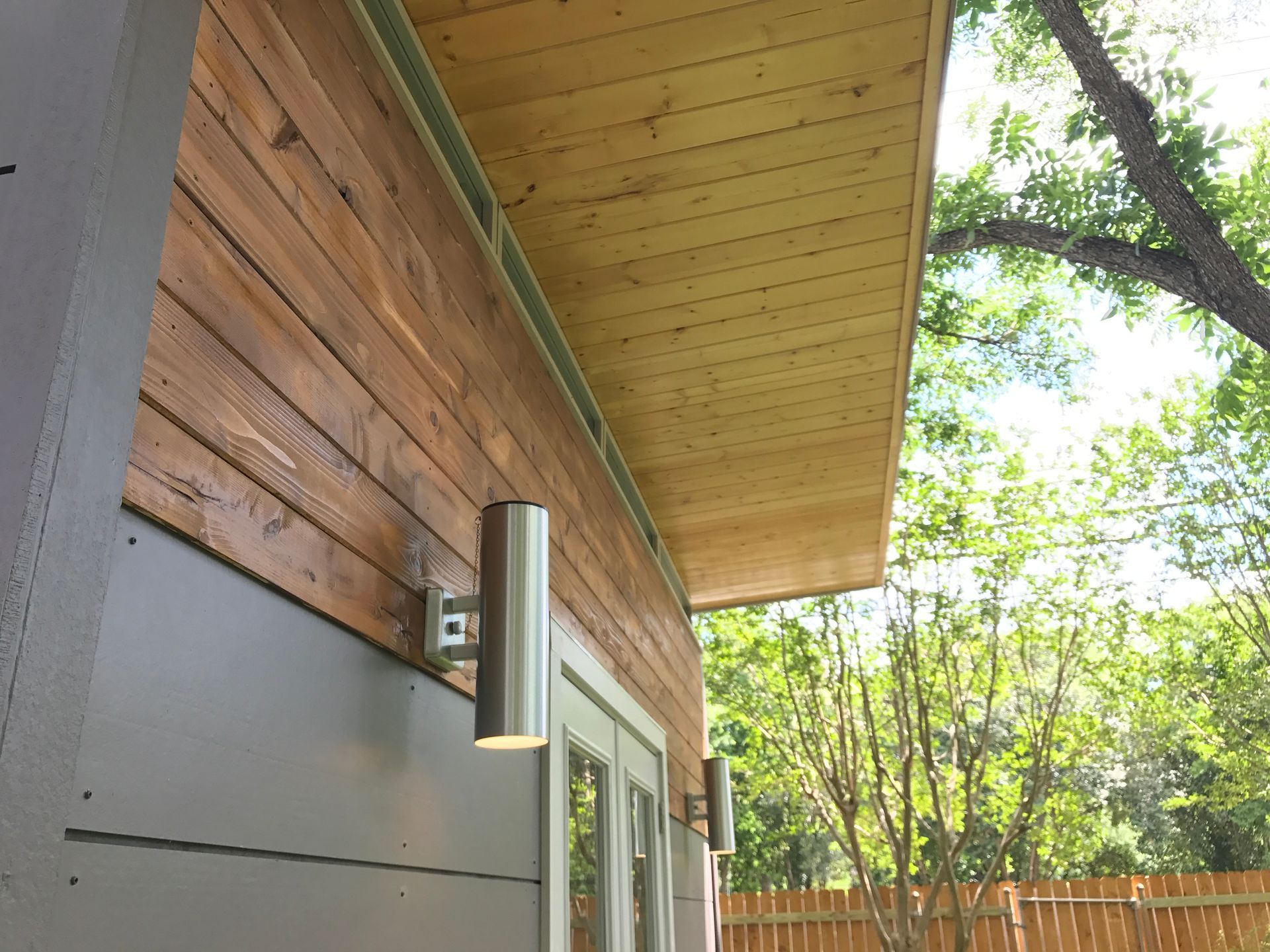 Exterior of a building with wooden siding, cylindrical lights, and a wooden ceiling under an overhang.