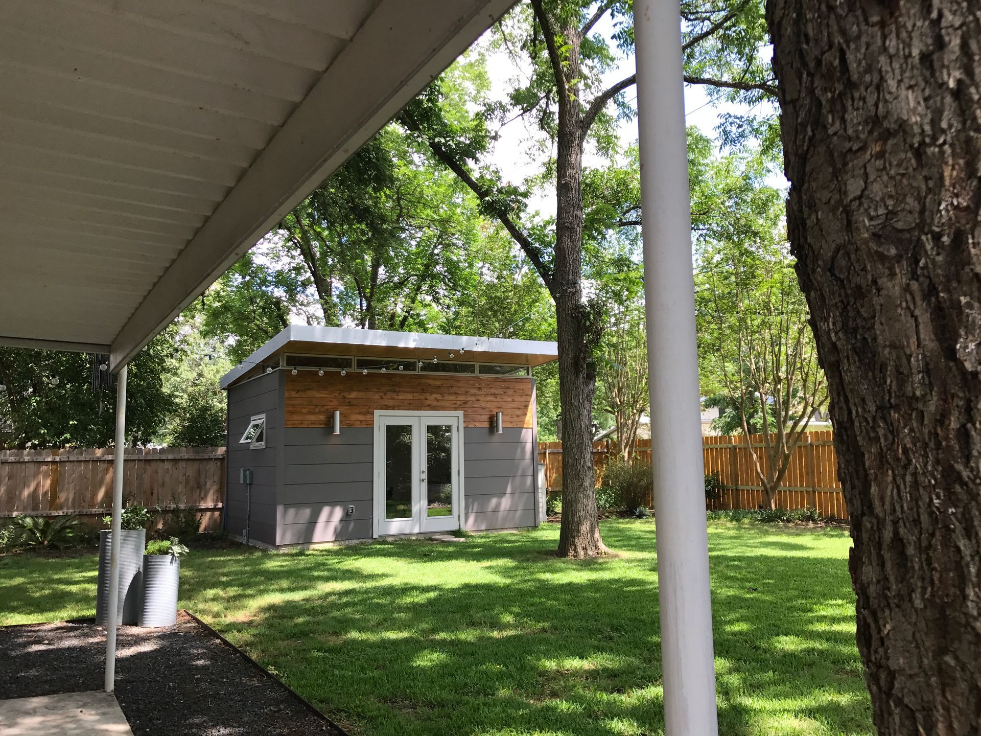 Backyard with a modern shed. Gray and wood exterior, white double doors. Green grass and trees surround it.
